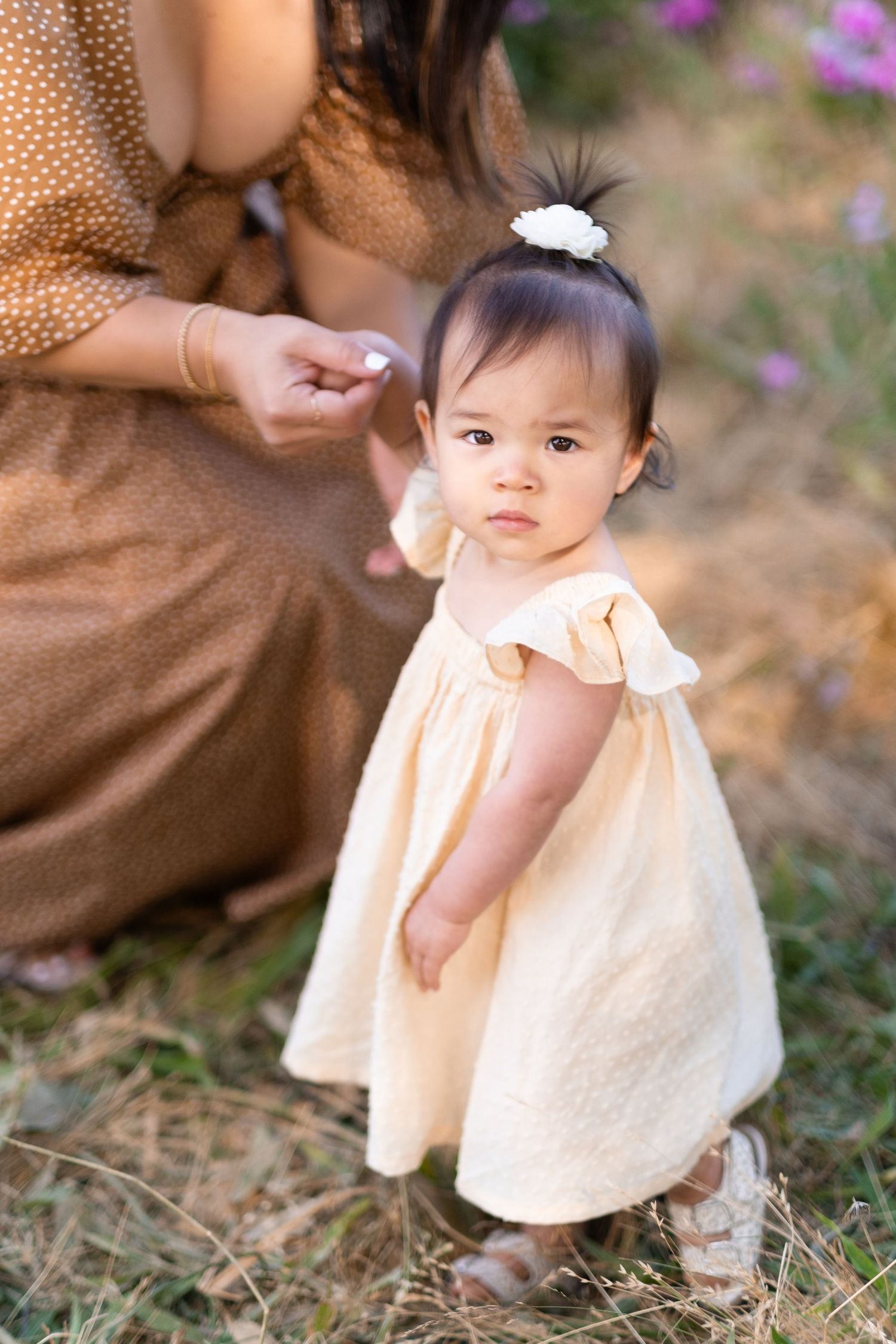 A small infant in a peach-colored dress standing in a garden while someone tends to them in brown attire.