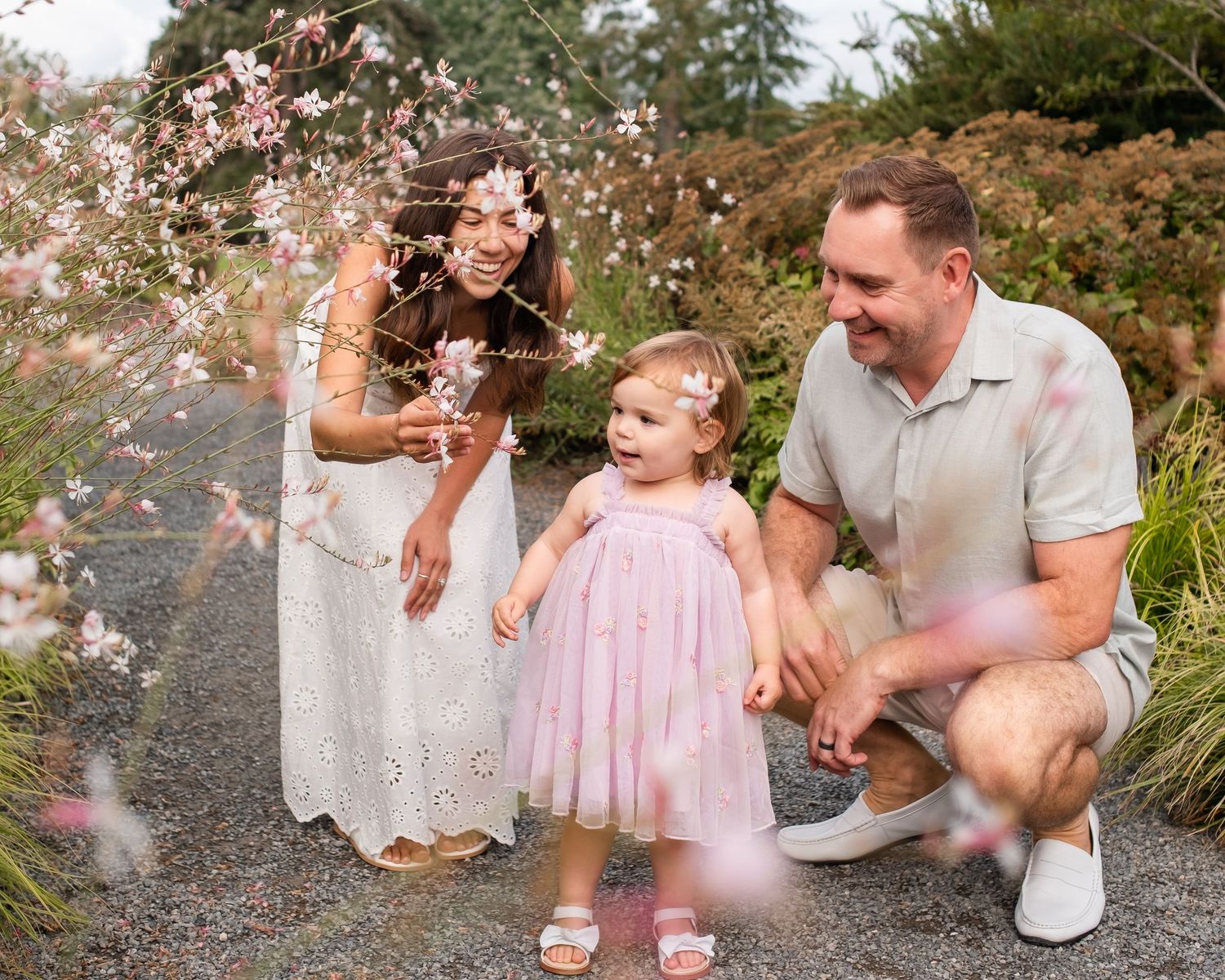 Family of three enjoying cherry blossoms in a garden, parents smiling at toddler in pink dress on a gravel path.