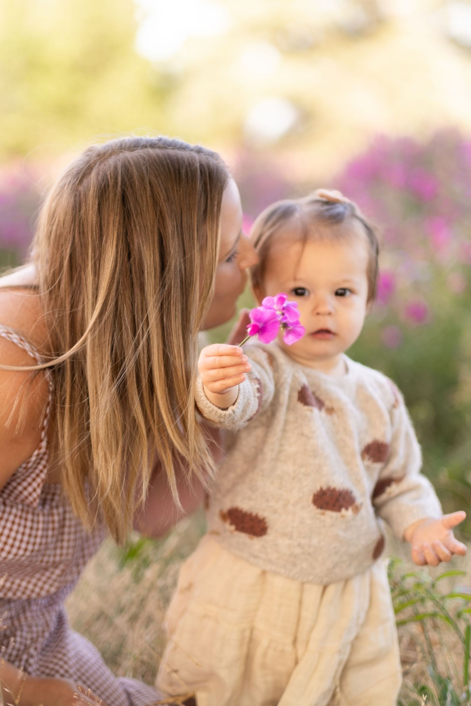 A toddler in a warm sweater holds a pink flower while standing with an adult in a garden setting with blooming flowers.
