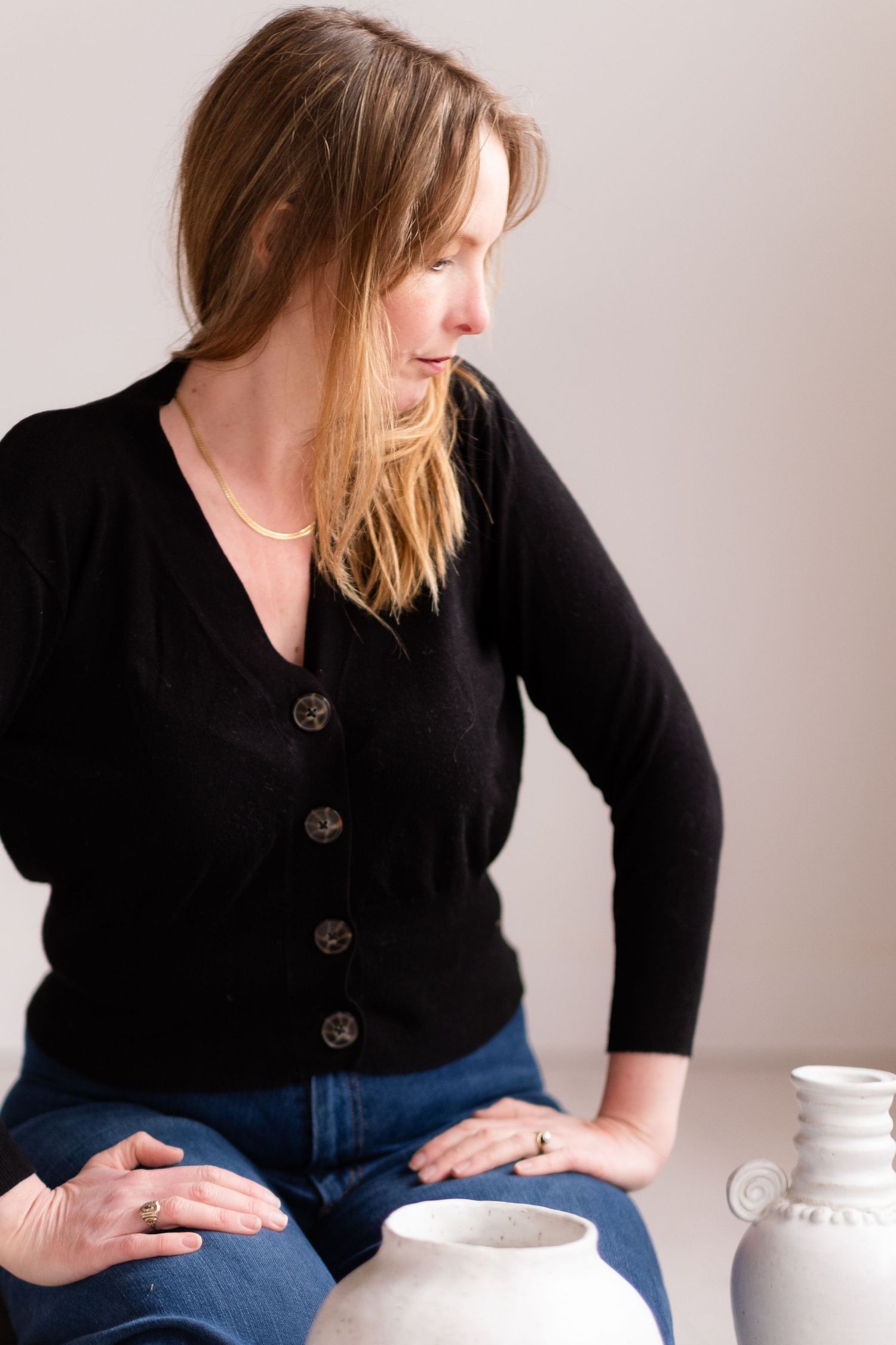 A potter in a black cardigan examines white ceramic pieces while seated in a minimalist studio setting.