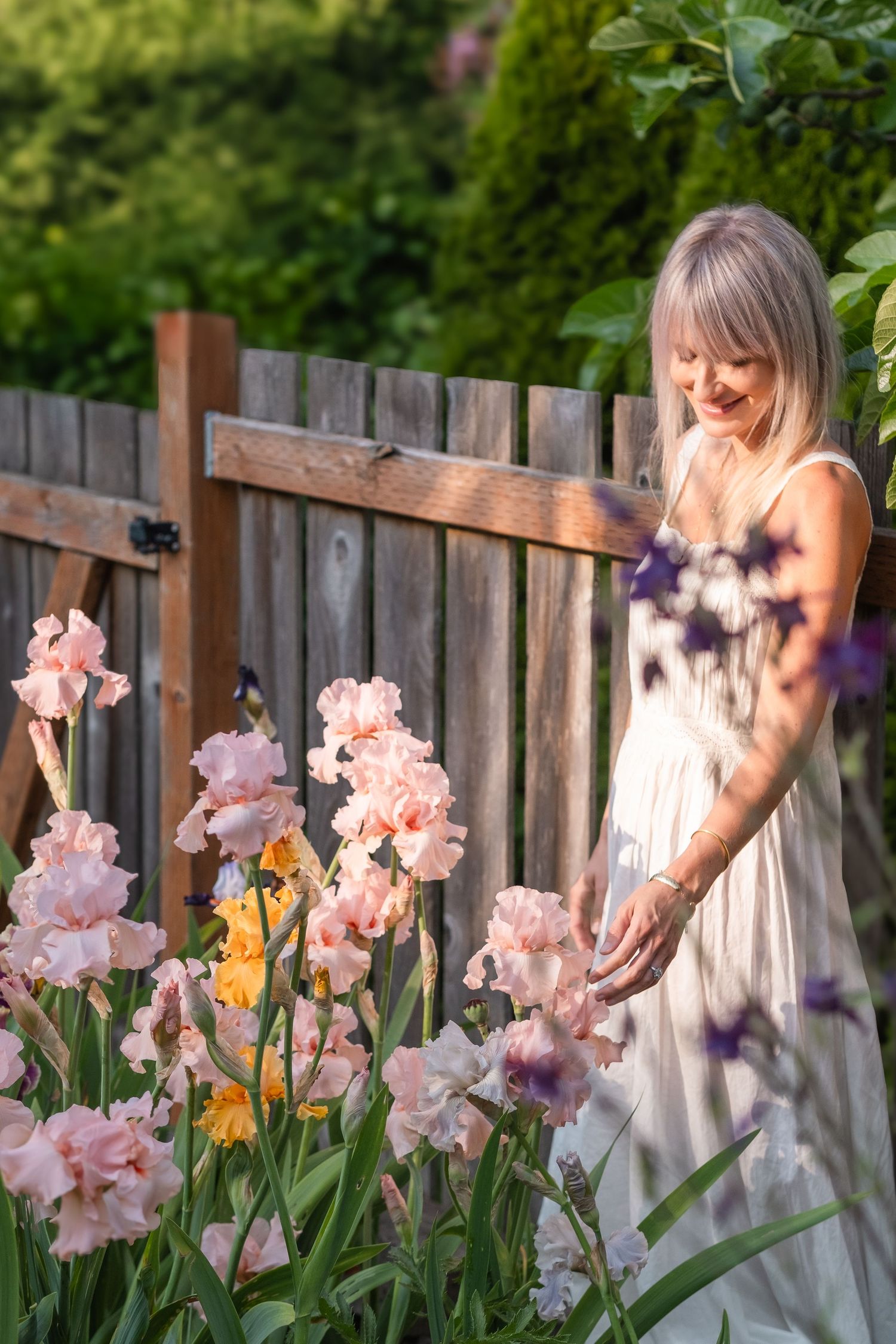 Smiling woman in white dress tending to pink and yellow iris garden.