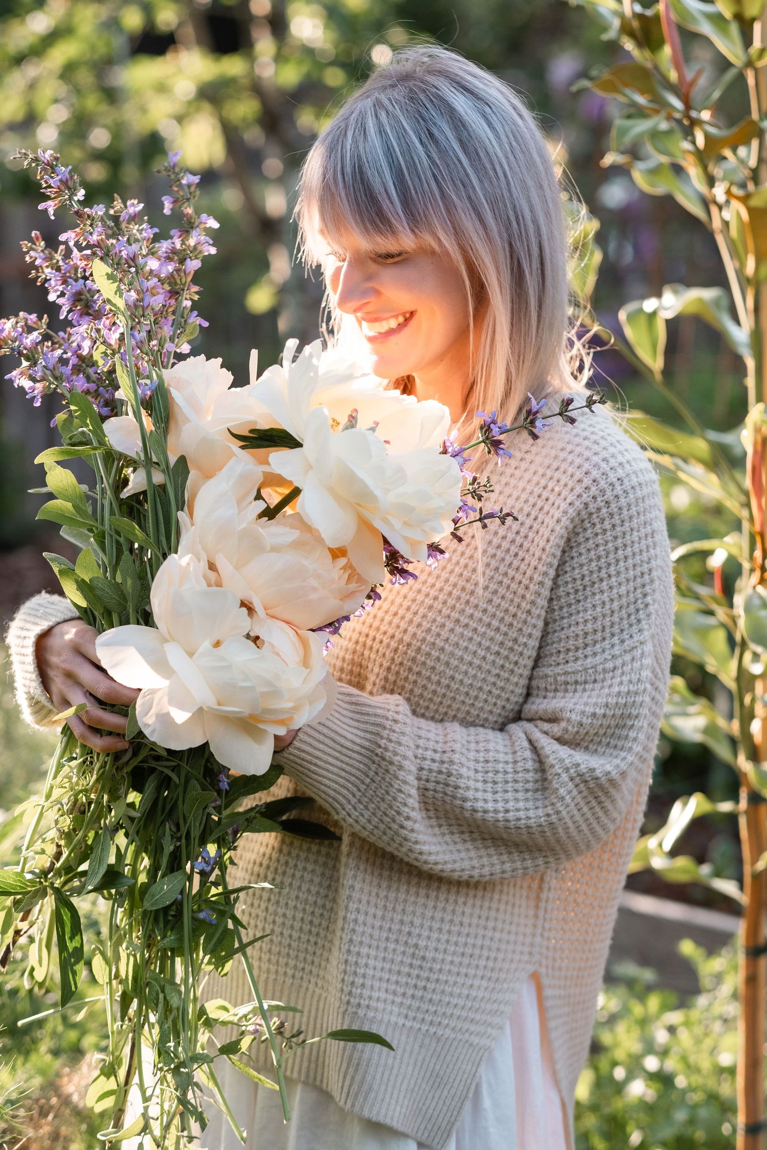 Happy blonde woman holding large white peonies and purple flowers in a sunny garden.