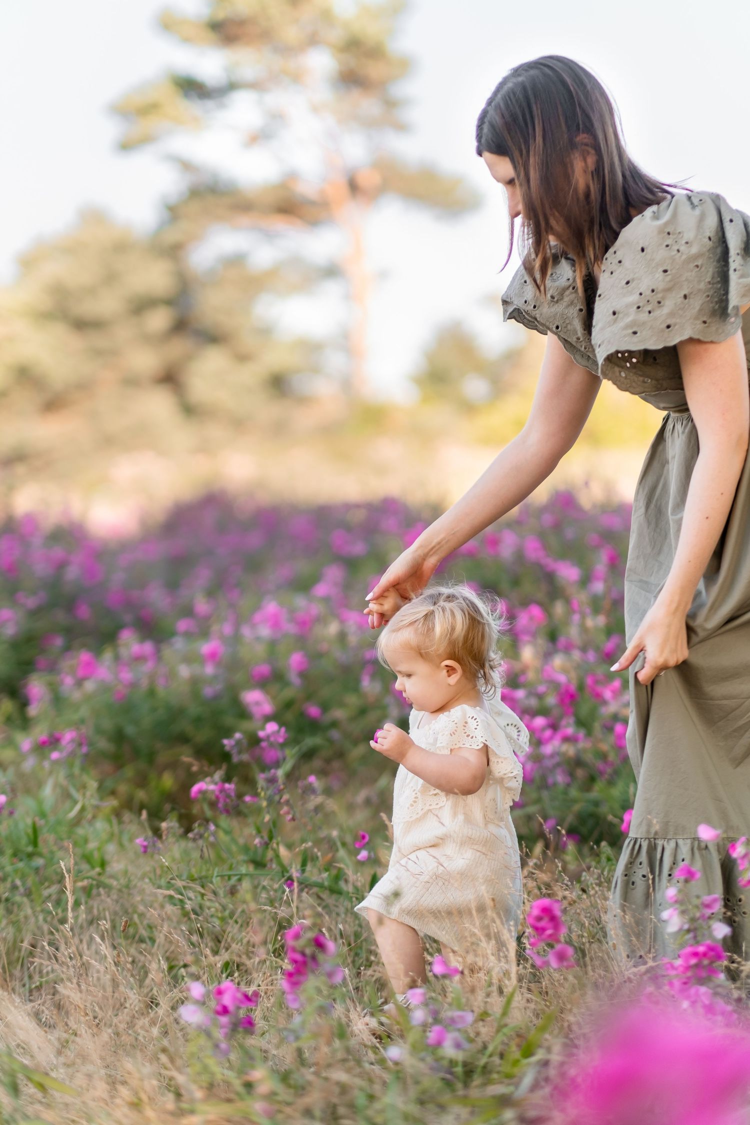 A figure in a sage dress guides a small child walking through blooming purple wildflowers in golden sunlight.