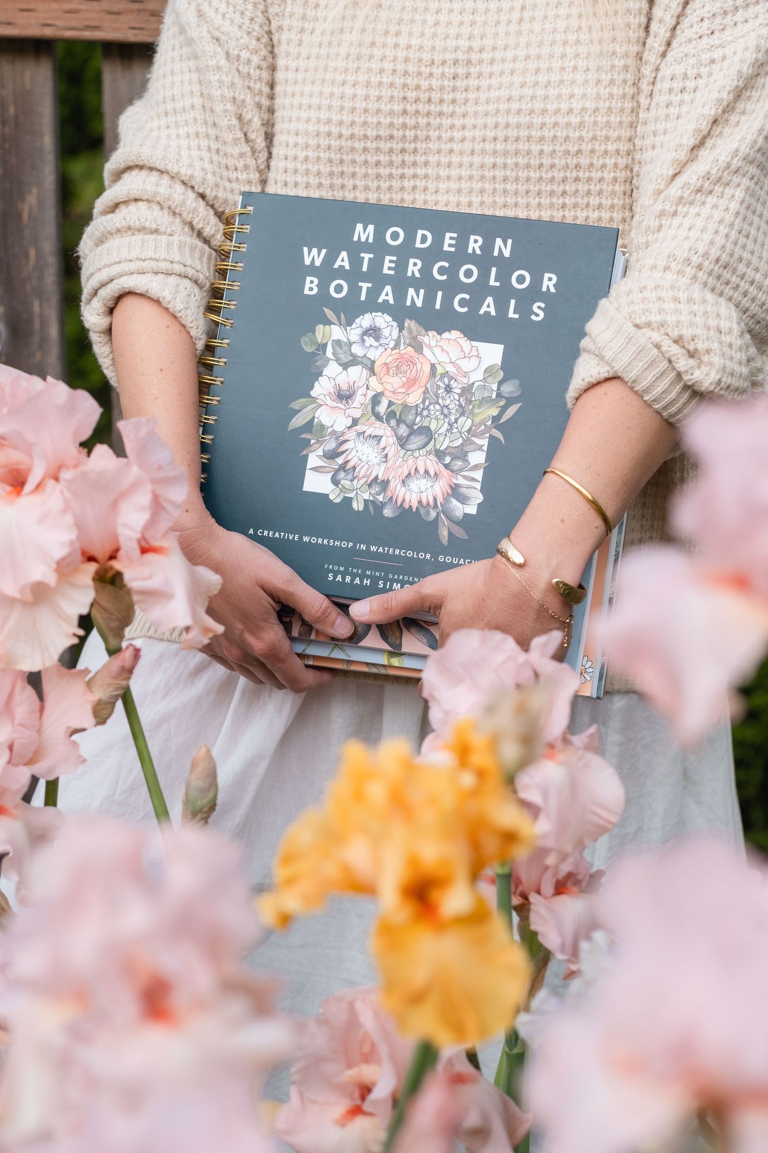 Person holding Modern Watercolor Botanicals spiral book surrounded by pink and yellow iris flowers in garden.