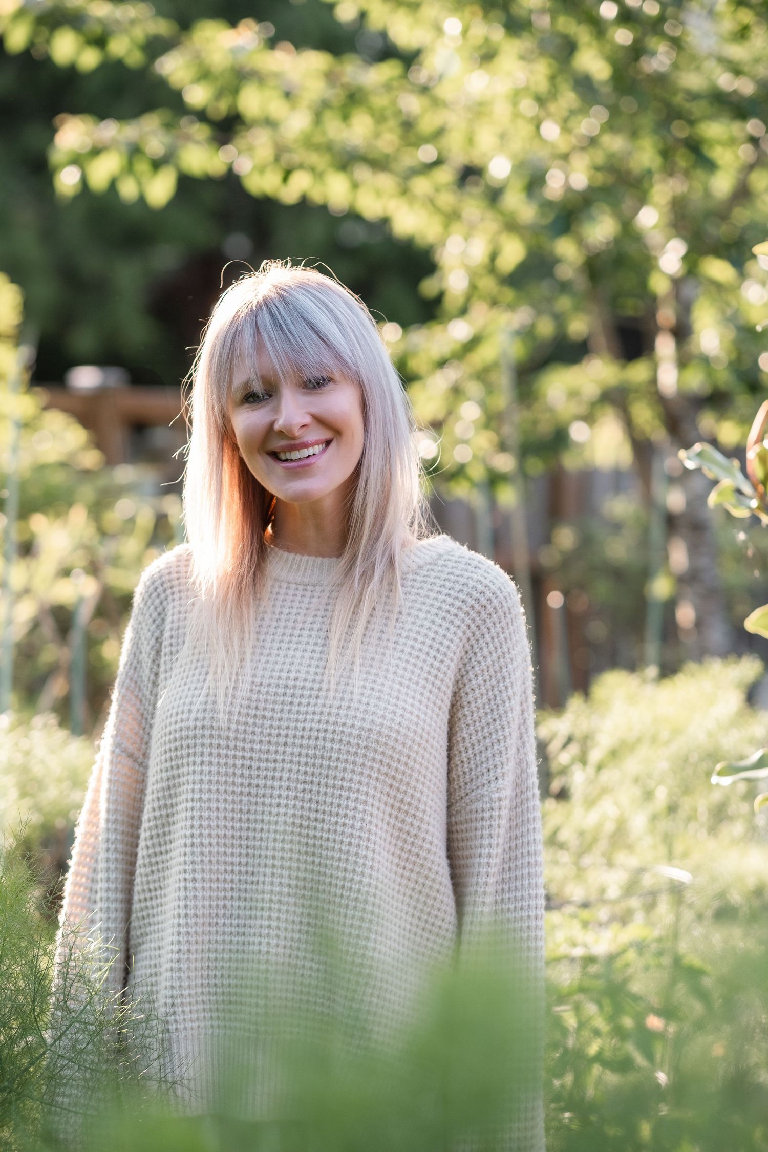 Smiling blonde woman in a cream knit sweater standing in a lush green garden.