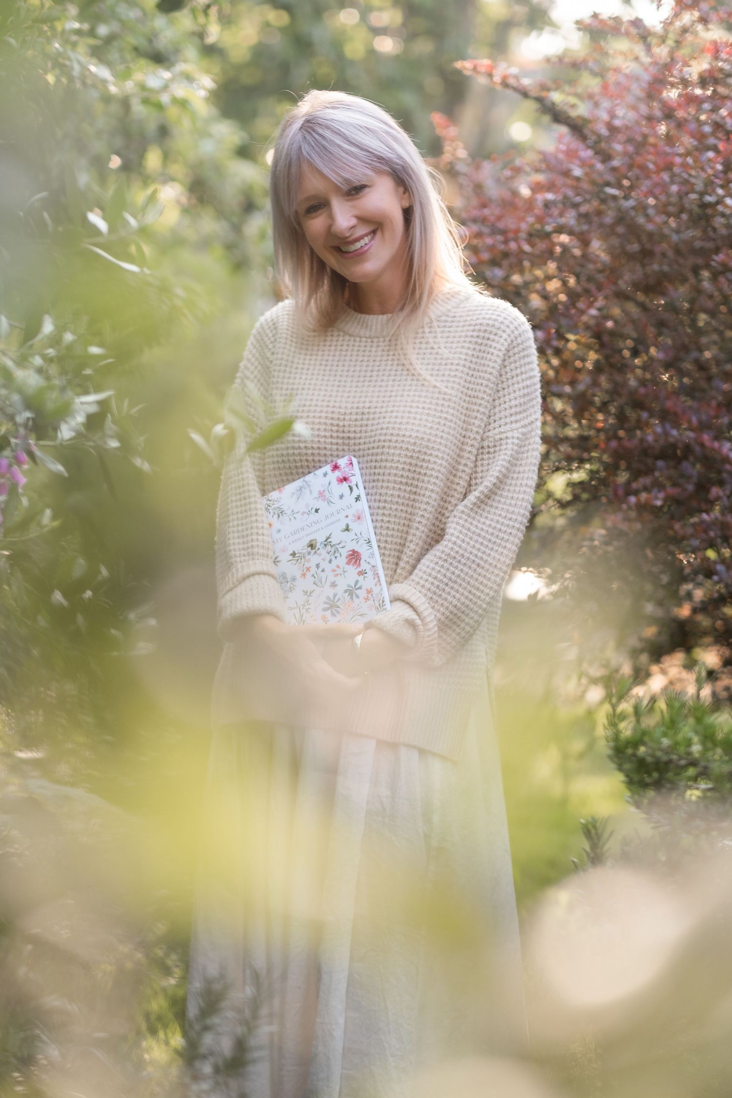 Smiling woman with blonde hair holding a floral book in a sunny garden.