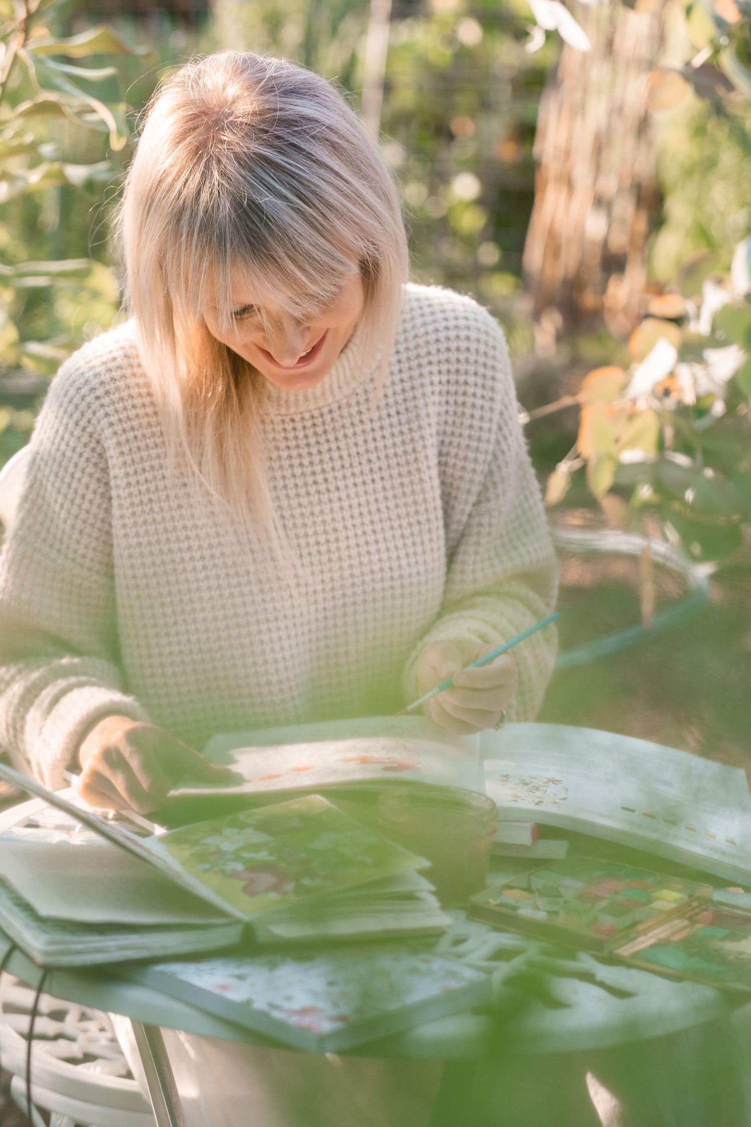 Blonde woman in cream waffle knit sweater reading a book outdoors in a sunlit garden.