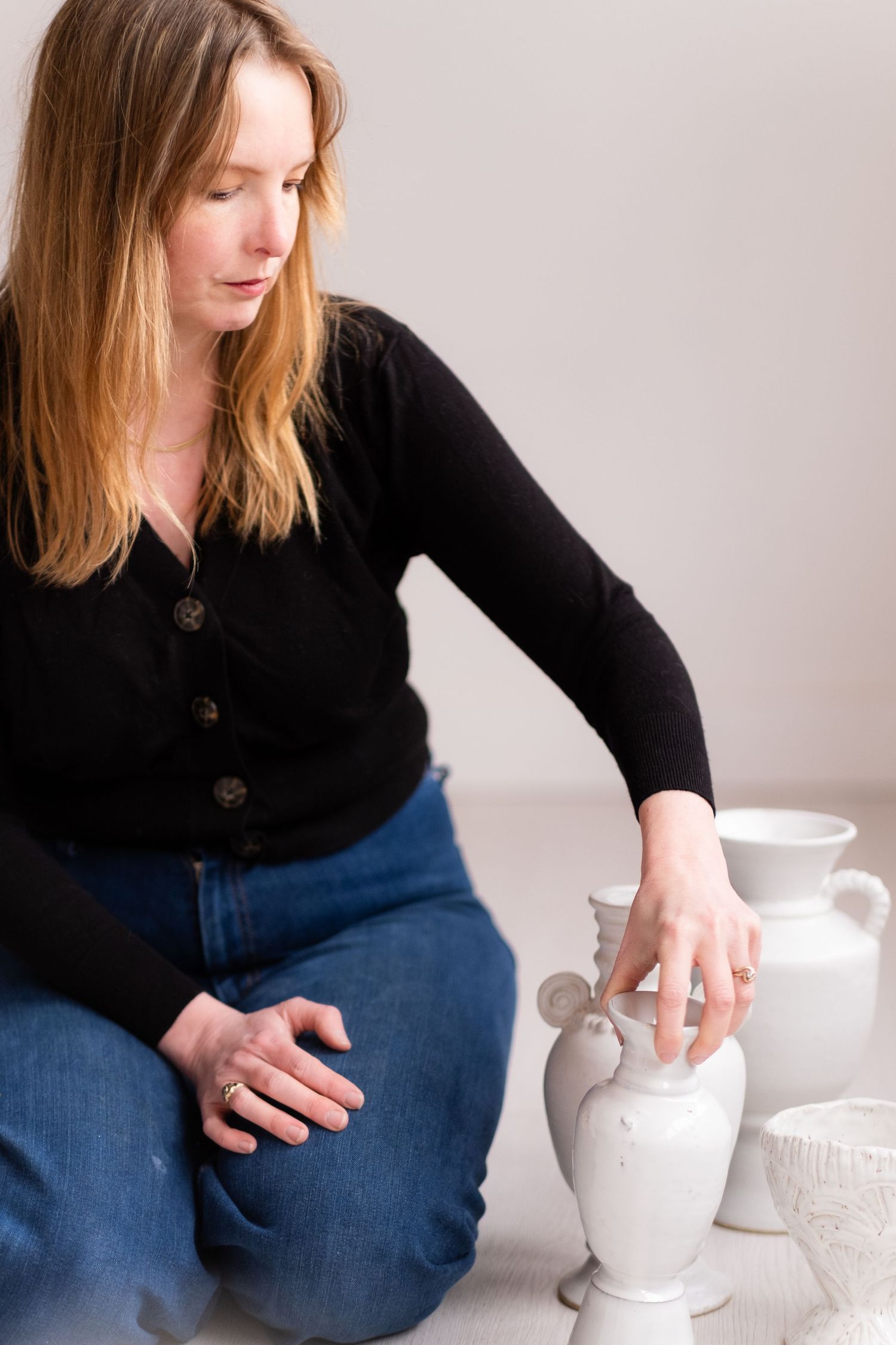 Person in black cardigan examining white ceramic vessels on light surface.
