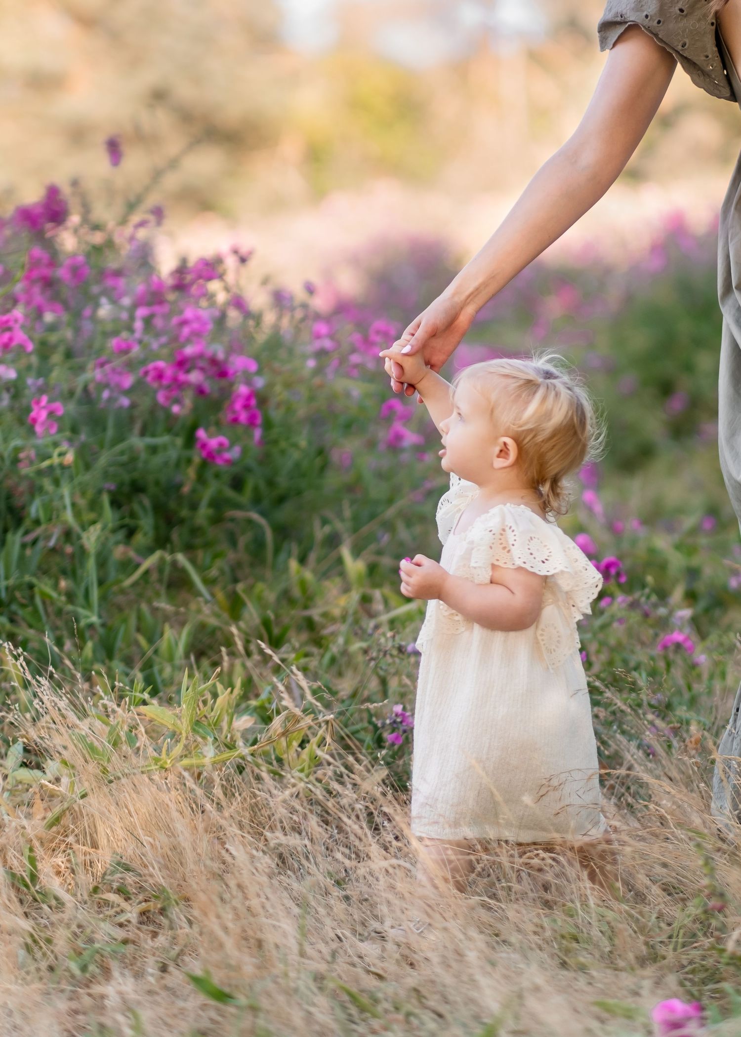 A small child in a white lace dress holds hands by purple wildflowers in a dreamy golden summer meadow.