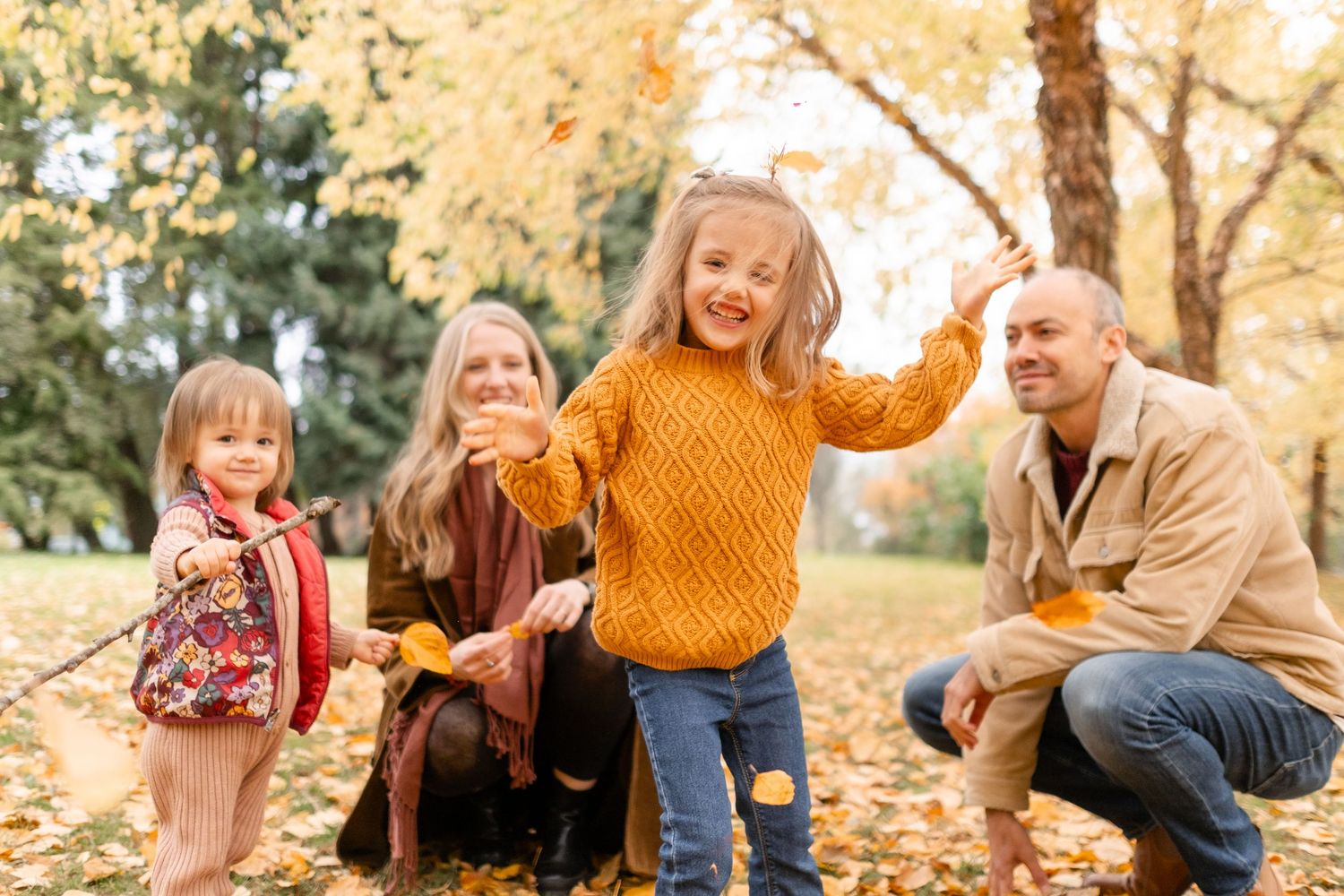 Family of four enjoying autumn park, parents and two young girls playing with fallen leaves among golden trees.