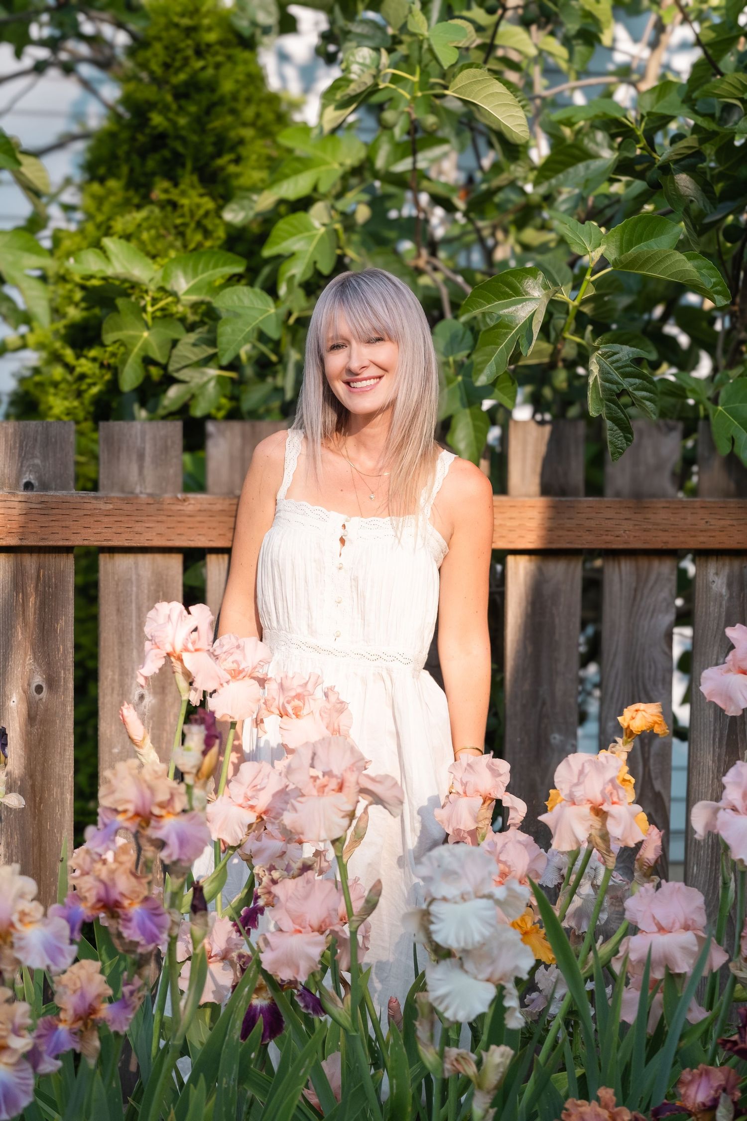 Smiling blonde woman in white sundress standing behind pink and purple iris flowers by a wooden garden fence.