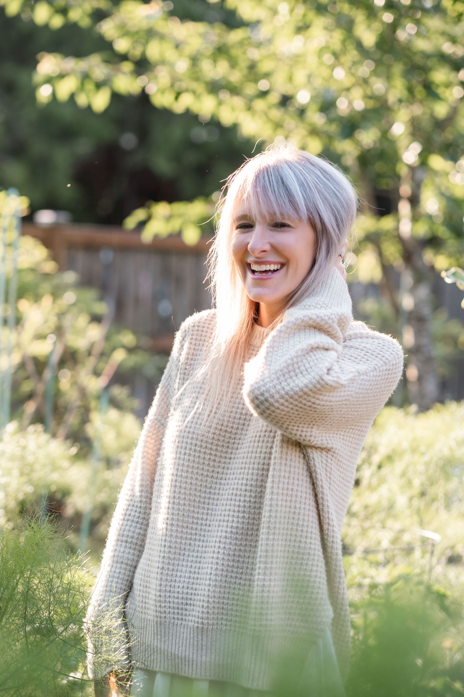 Smiling blonde woman in cream waffle knit sweater laughing outdoors in a lush green garden.