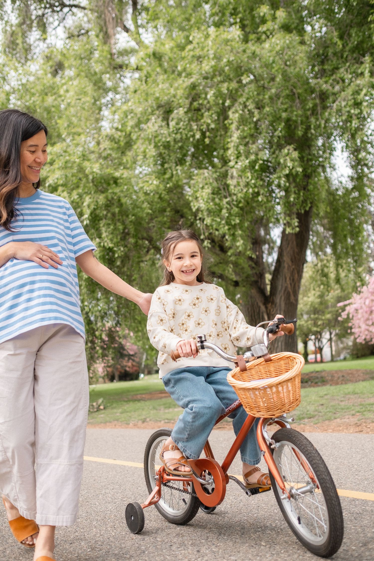 A child learns to ride an orange bicycle in a park while an adult provides guidance and support.