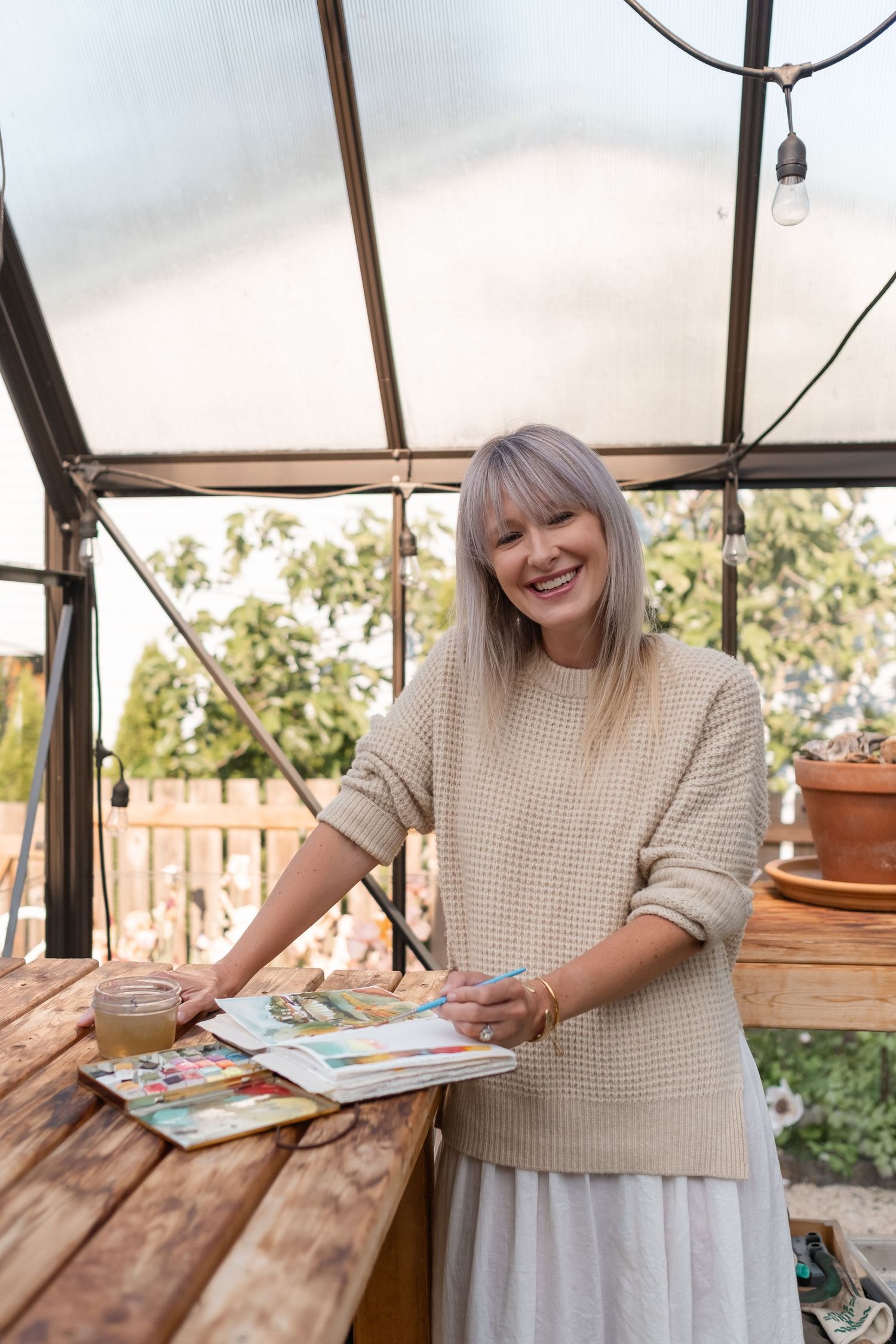 Happy blonde woman in beige sweater holding watercolor sketchbook in a sunlit greenhouse studio.