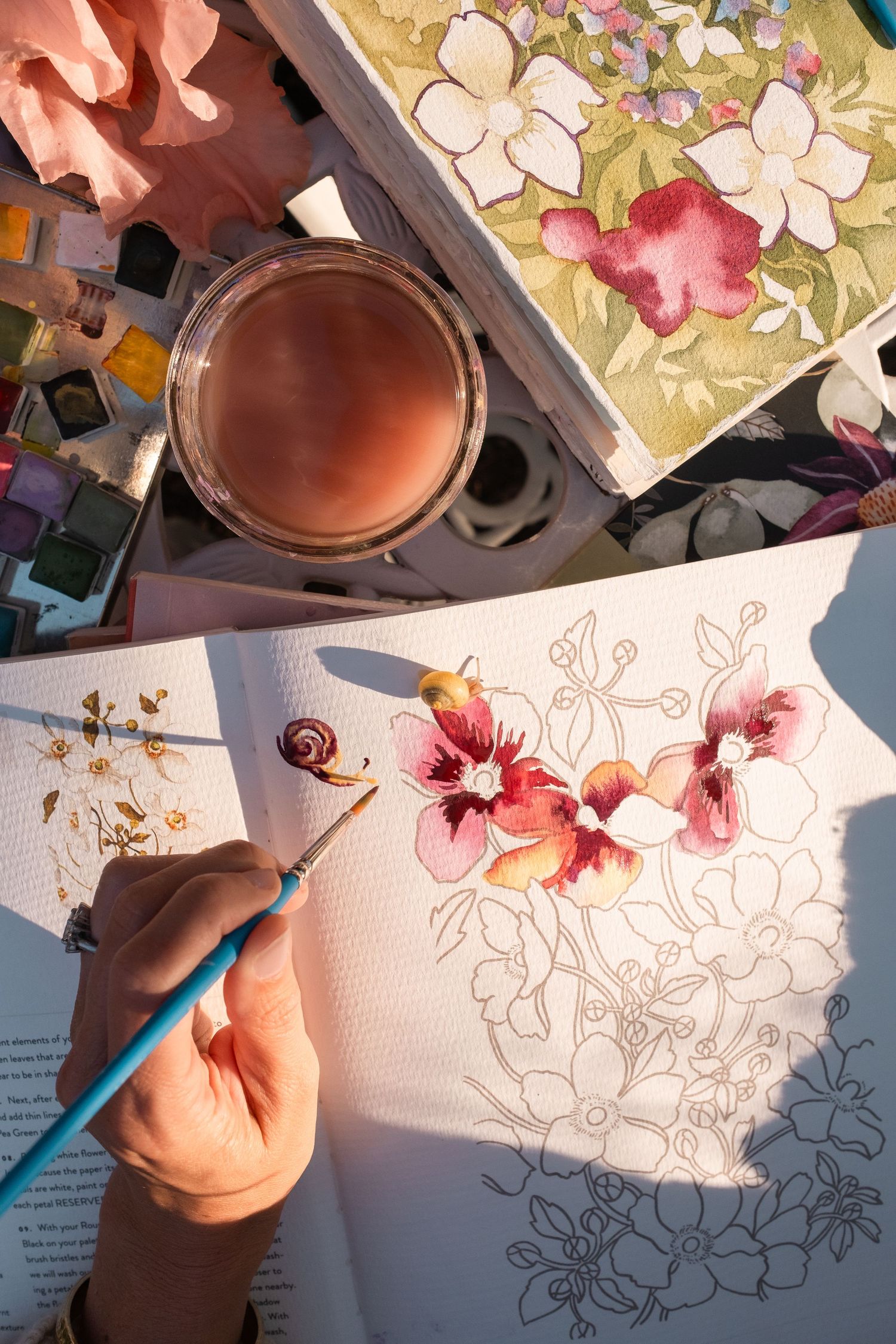 Close-up of hands painting colorful floral watercolor artwork with brushes and paints on a table.