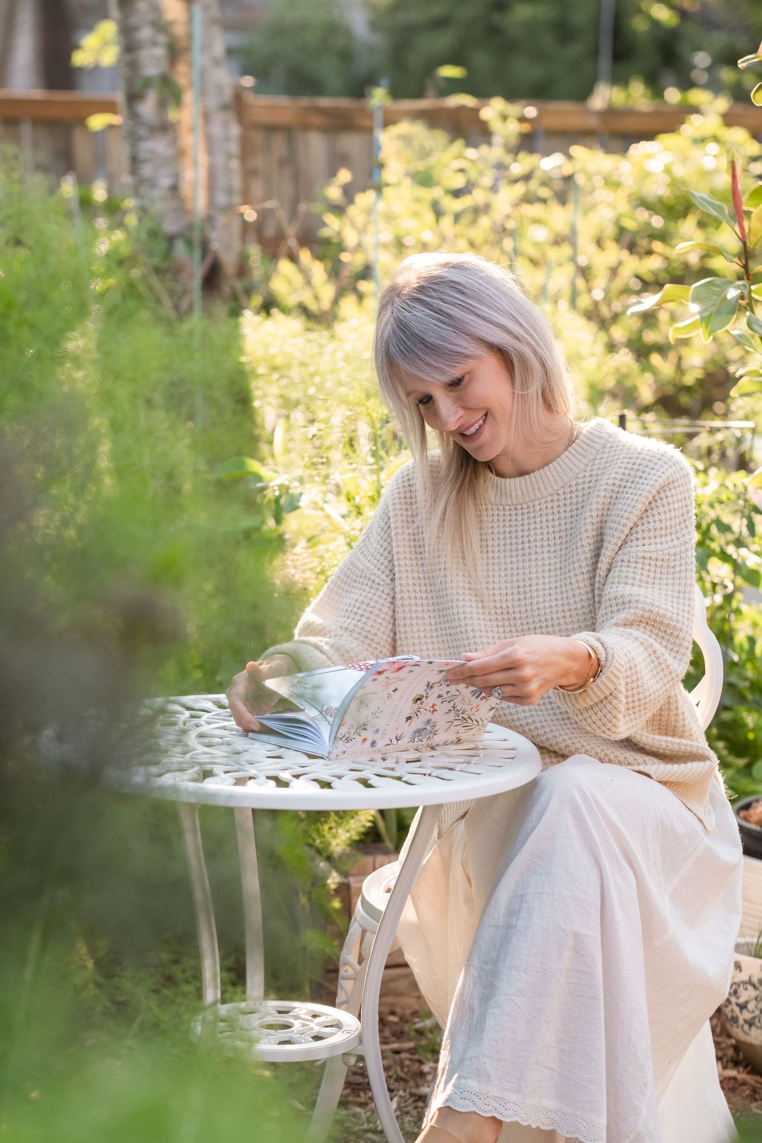 Happy blonde woman in beige sweater reading a floral magazine at a white garden table.