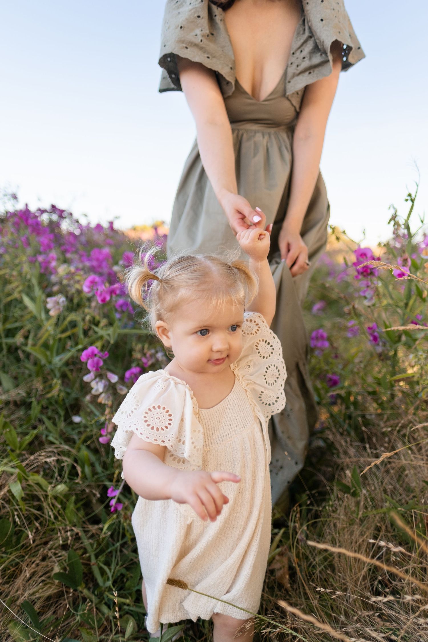 A toddler in a lace-trimmed dress stands among purple wildflowers while being guided by someone in a sage green dress.