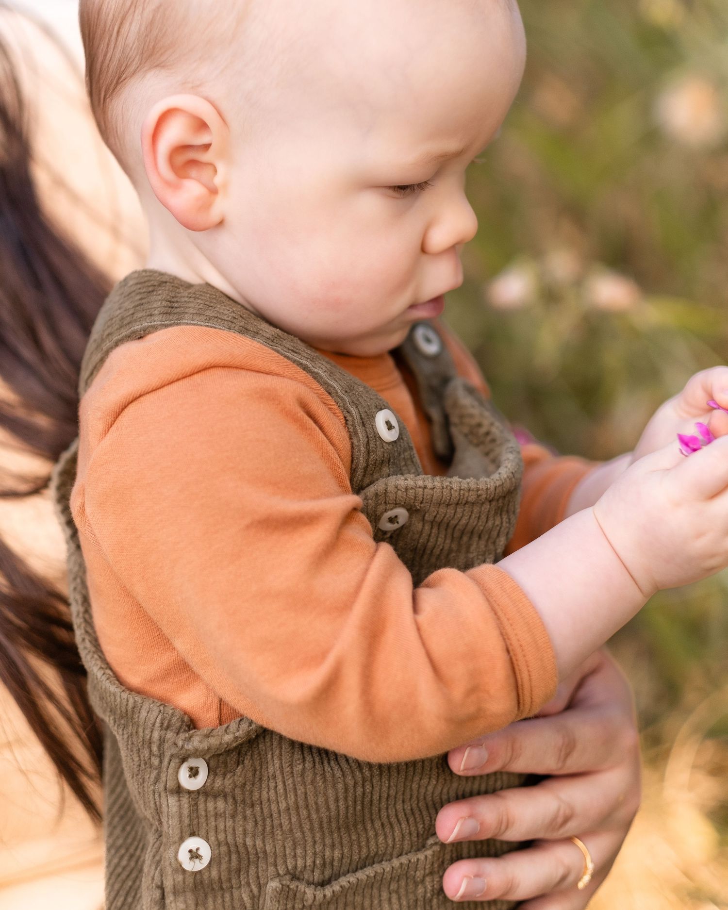 Baby in orange sweater and brown corduroy overalls being held while examining something in their hands outdoors.