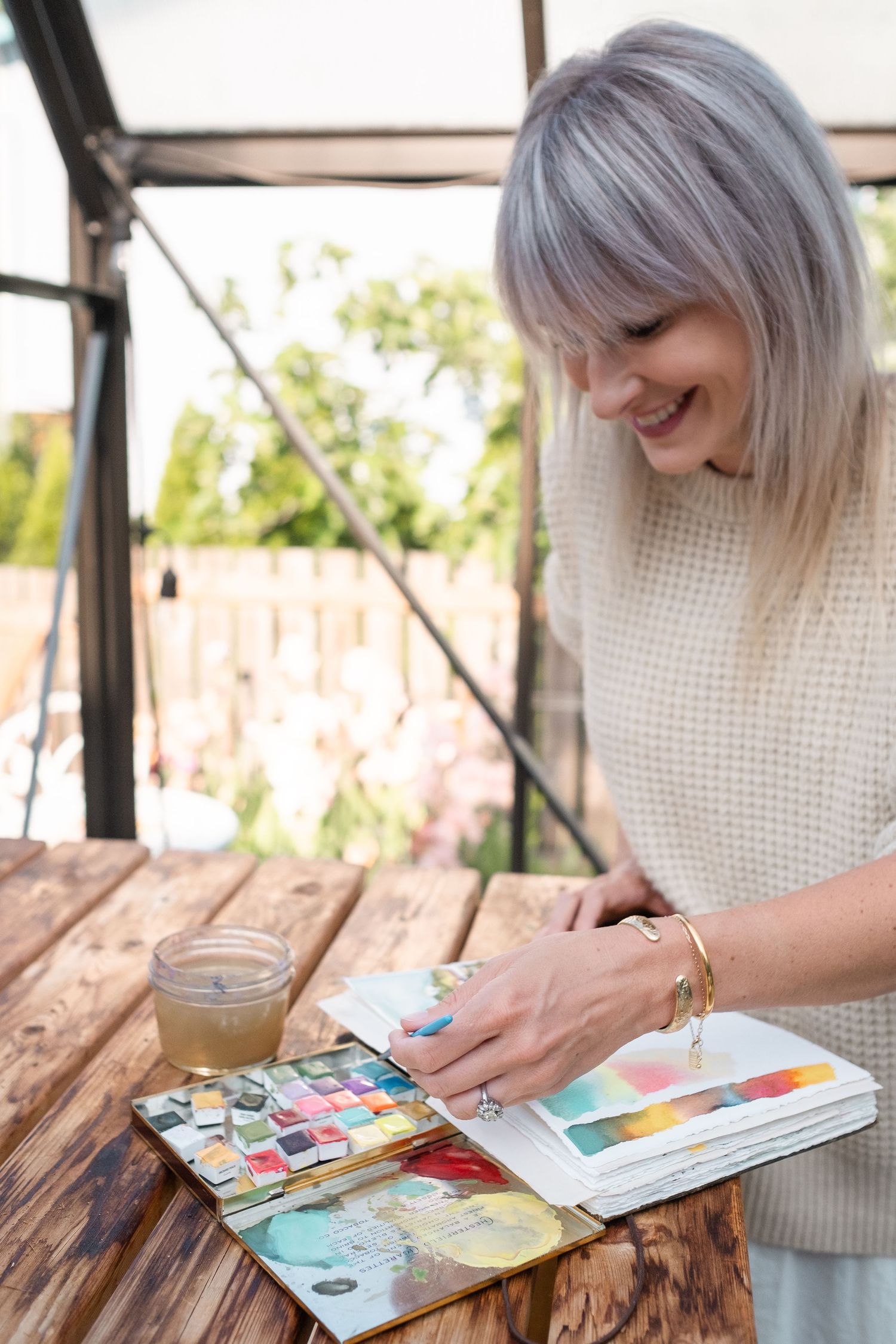 Smiling woman with silver hair reviewing colorful watercolor sketchbook on wooden table in glass greenhouse studio.