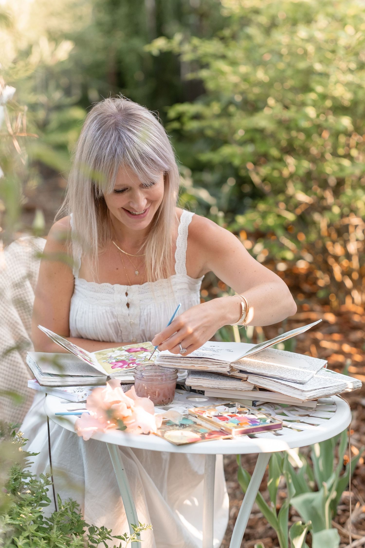 Smiling blonde woman painting watercolors in an open sketchbook at a white outdoor garden table.