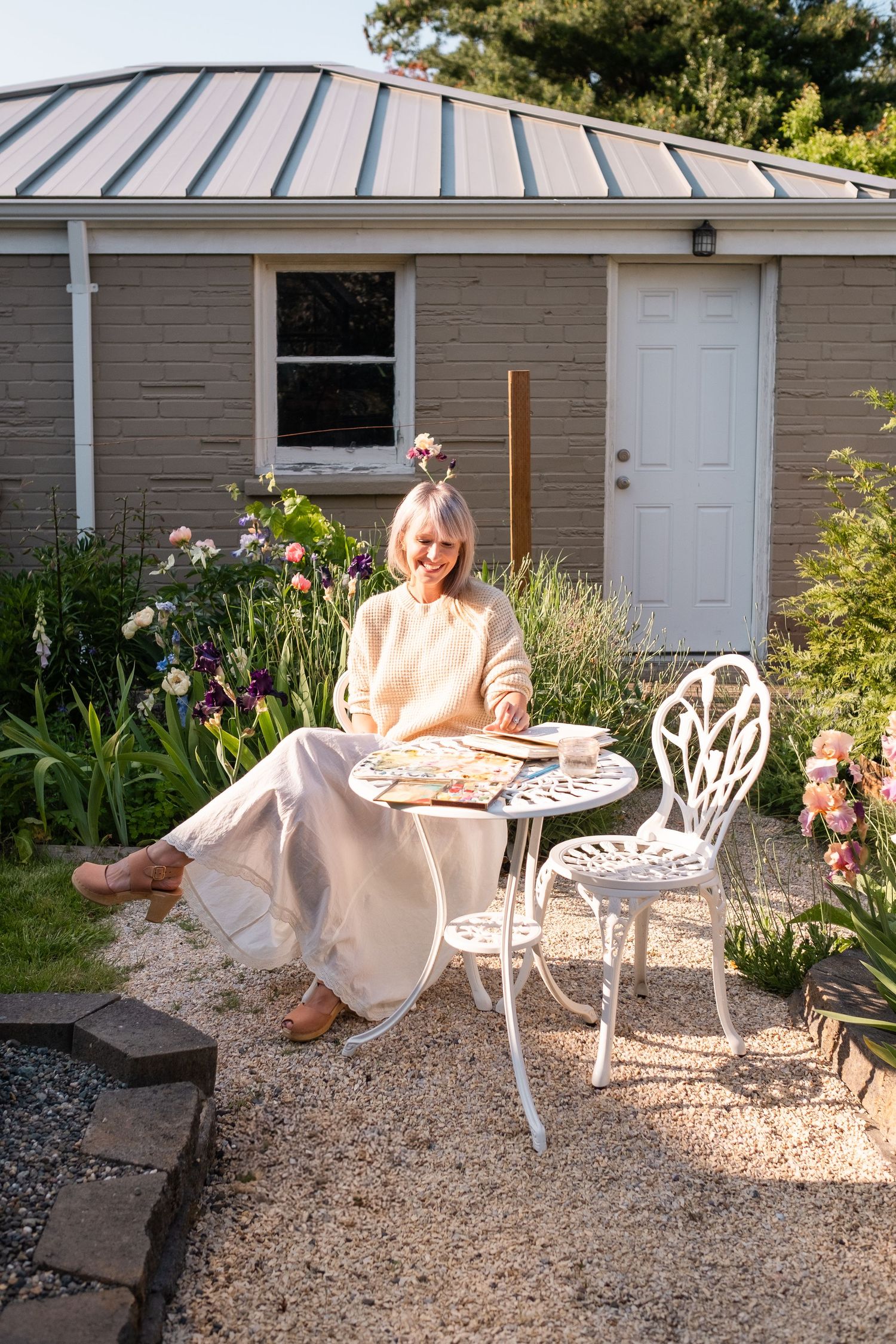 Woman in cream outfit relaxing at ornate white metal garden table with watercolor sketchbook in sunny backyard.