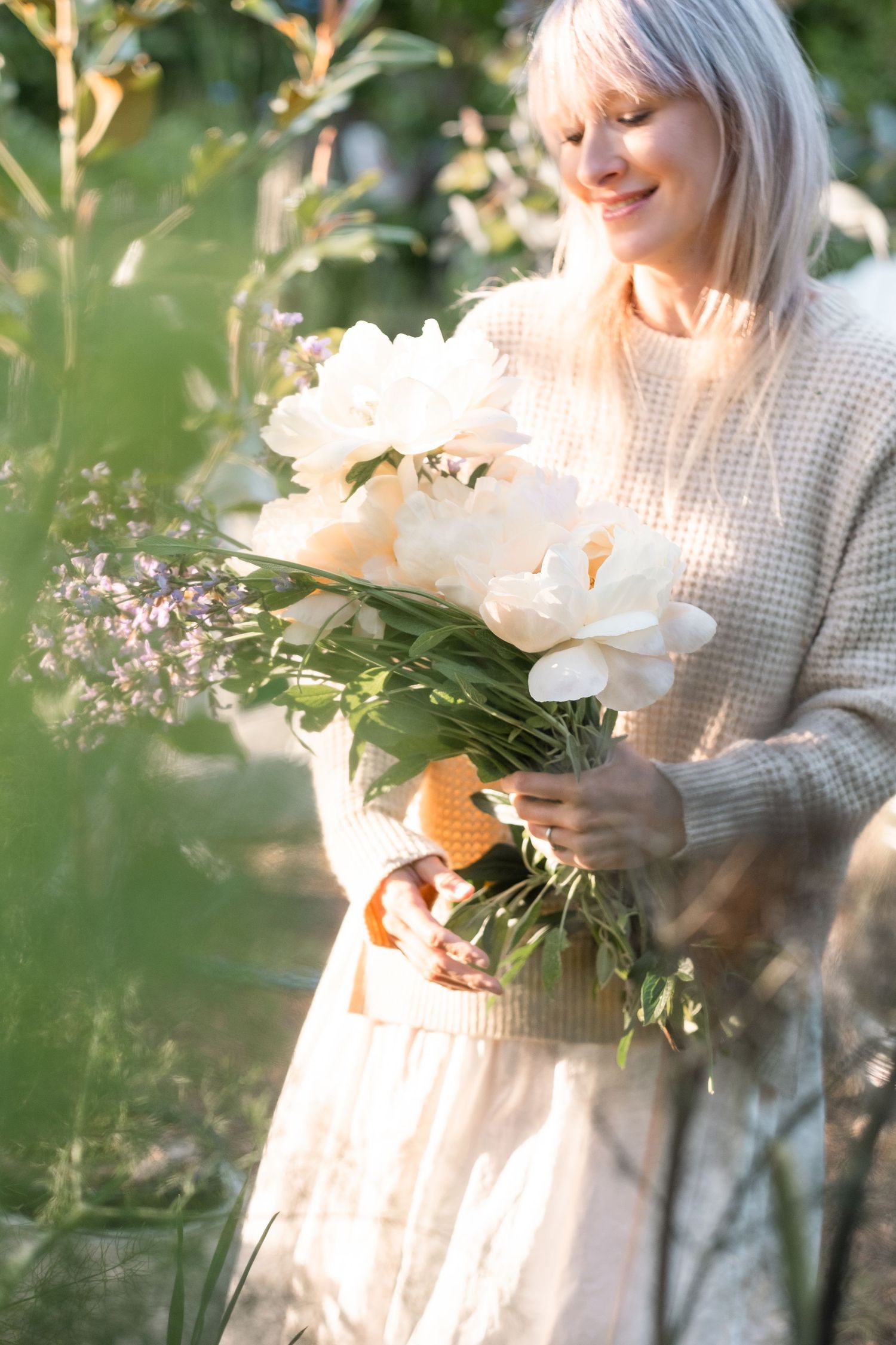 Smiling blonde woman holding a bouquet of white peonies and wildflowers outdoors in soft golden sunlight.