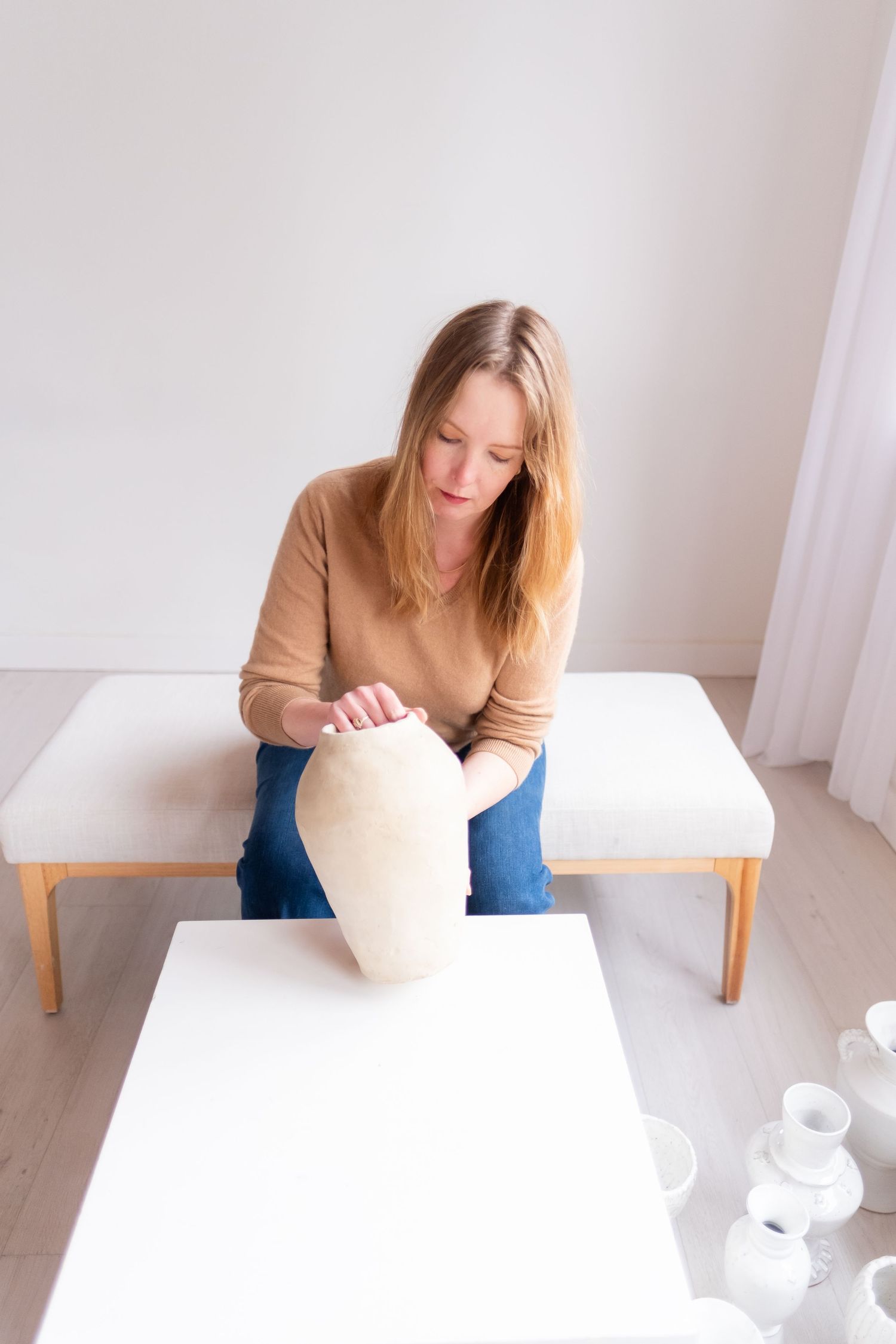 Artist works on white ceramic piece while seated at table in bright minimal studio space.