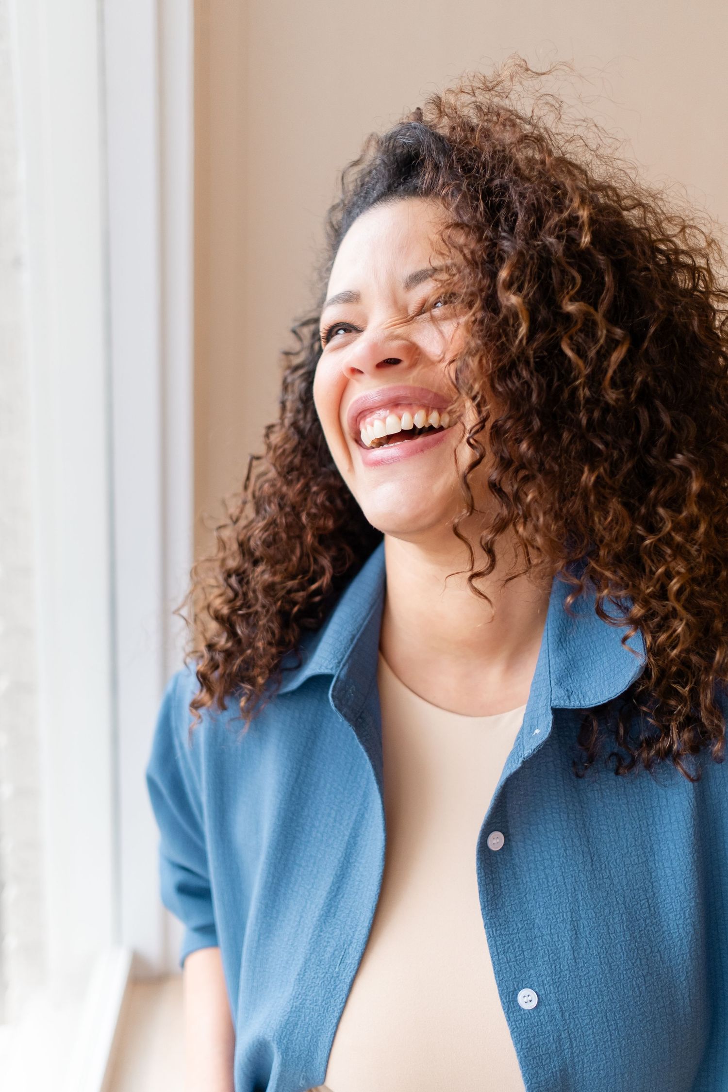 A person with curly brown hair wearing a blue button-up shirt laughs joyfully near a window.