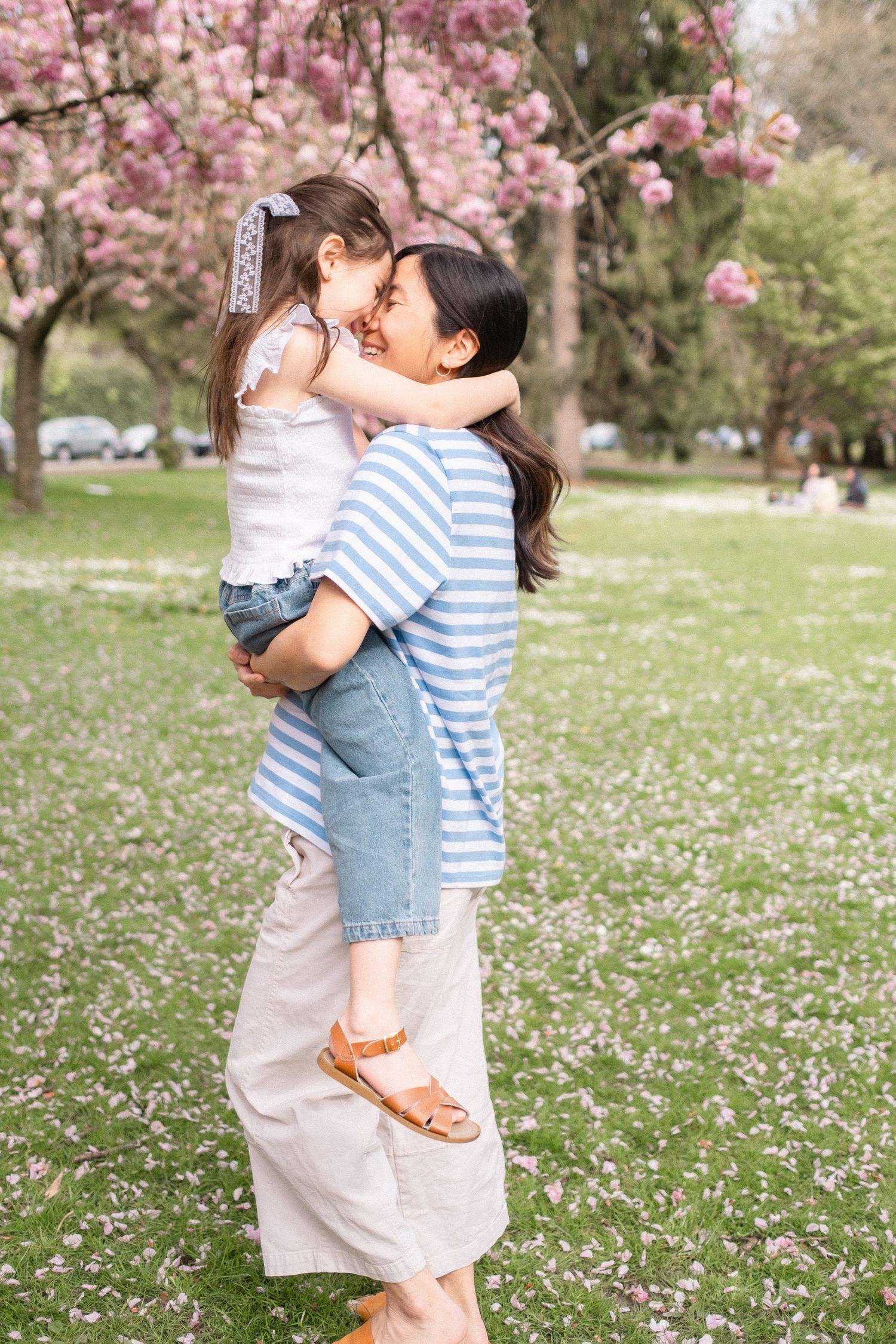 Two people embrace in a tender moment under blooming cherry blossoms while spring petals scatter on the grass.