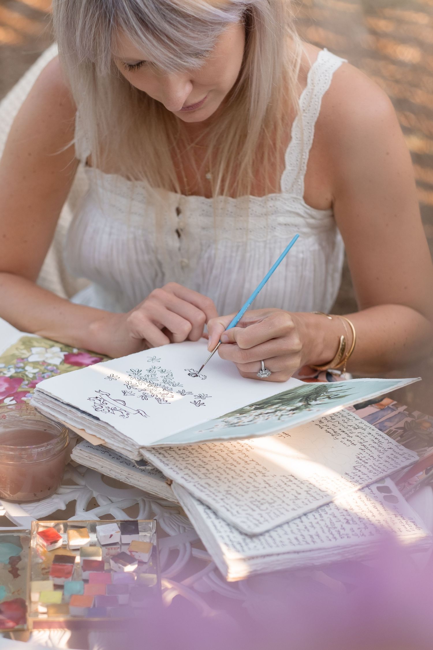 Blonde woman in white dress painting in sketchbook with blue brush at outdoor garden table.