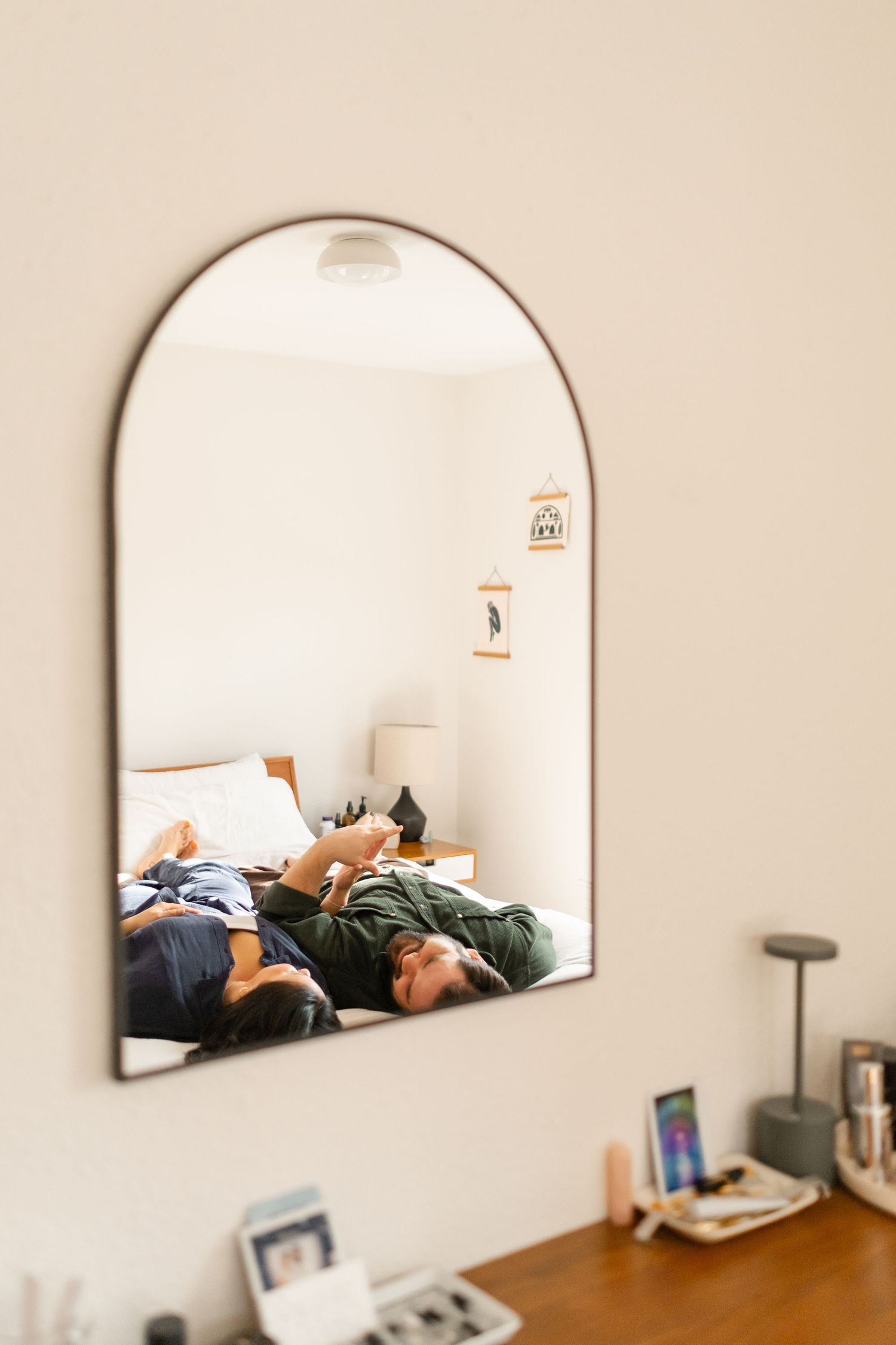 Mirror reflection shows couple relaxing on bed with phones while natural light streams into minimalist bedroom.