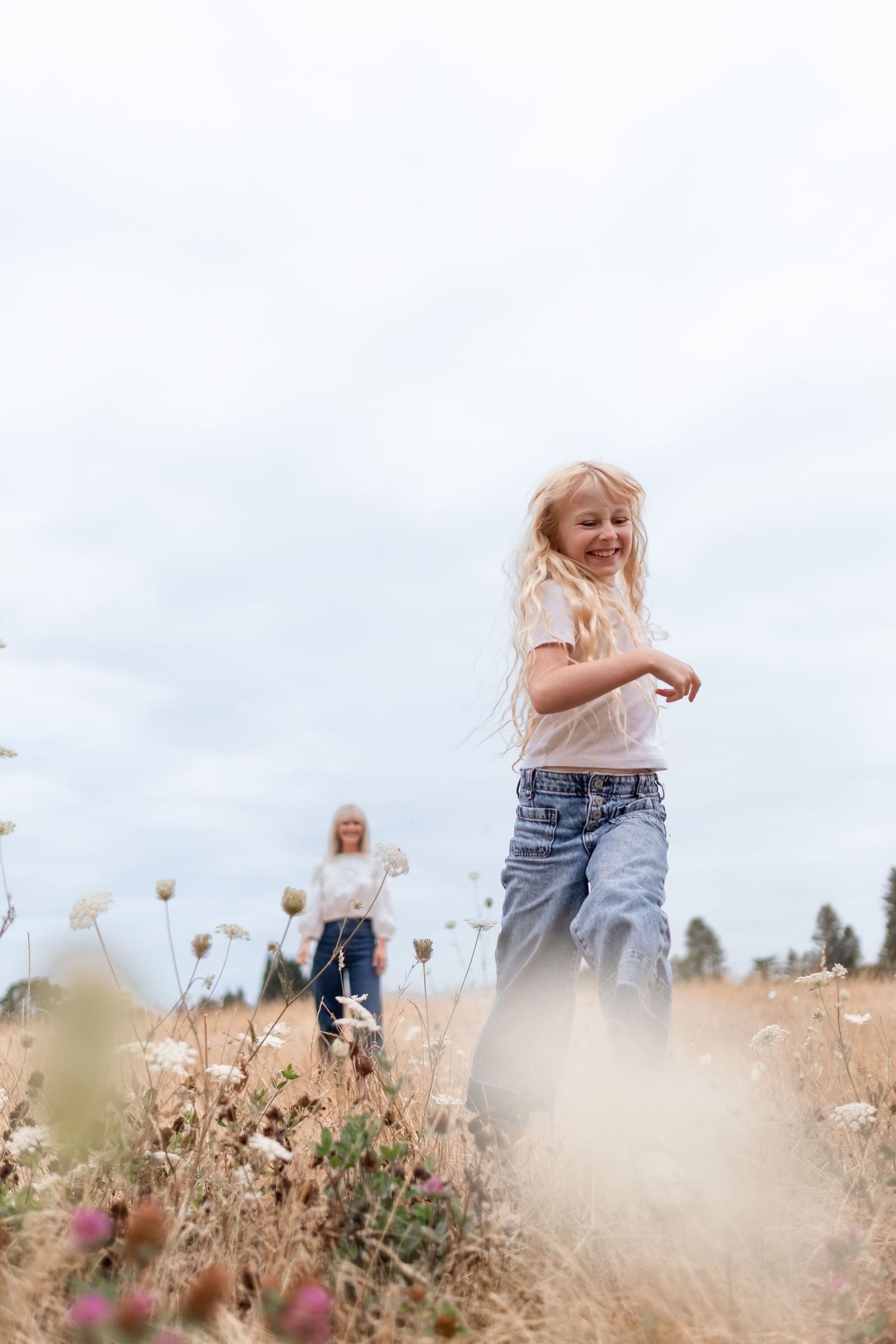A joyful blonde girl runs through a wildflower field while a woman stands in the background on a cloudy day.
