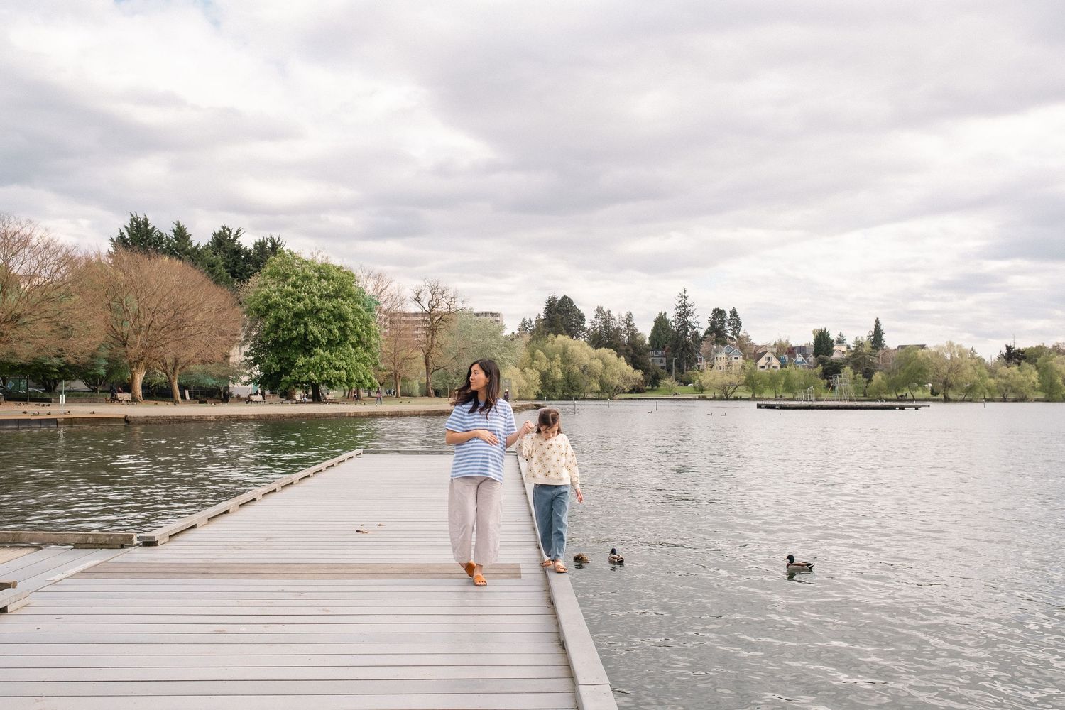 A couple walks their dog along a waterfront path on an overcast day with scenic views of the lake and trees.