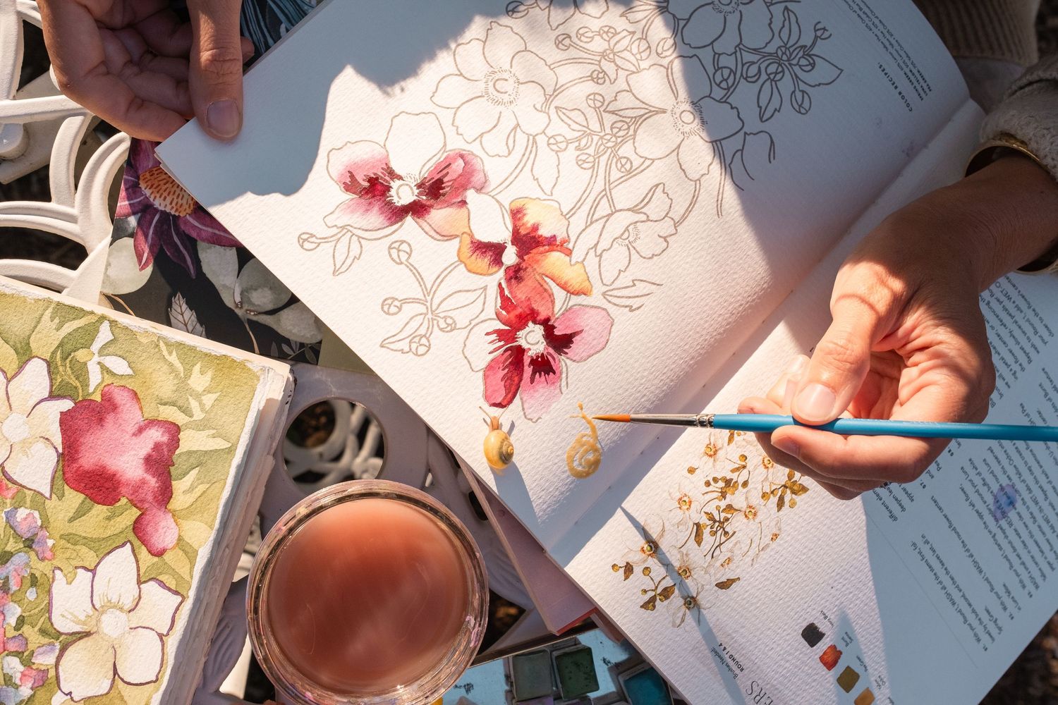 Overhead view of hands painting pink orchid watercolor with brushes and paint palette on table.