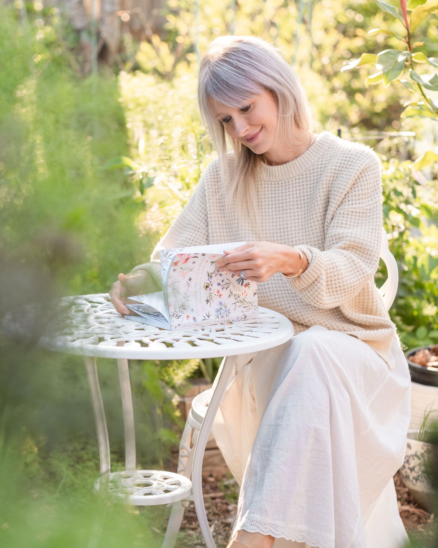 Woman in beige sweater sitting at white garden table opening a floral gift box outdoors.