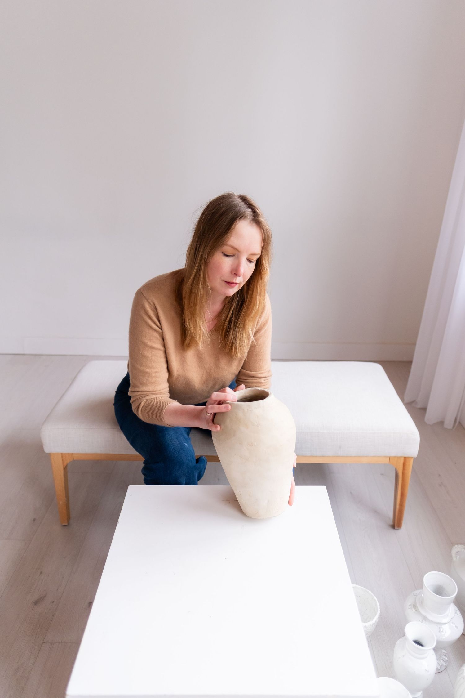 An artist studies a large white ceramic vase while seated on a wooden bench in a minimal setting.