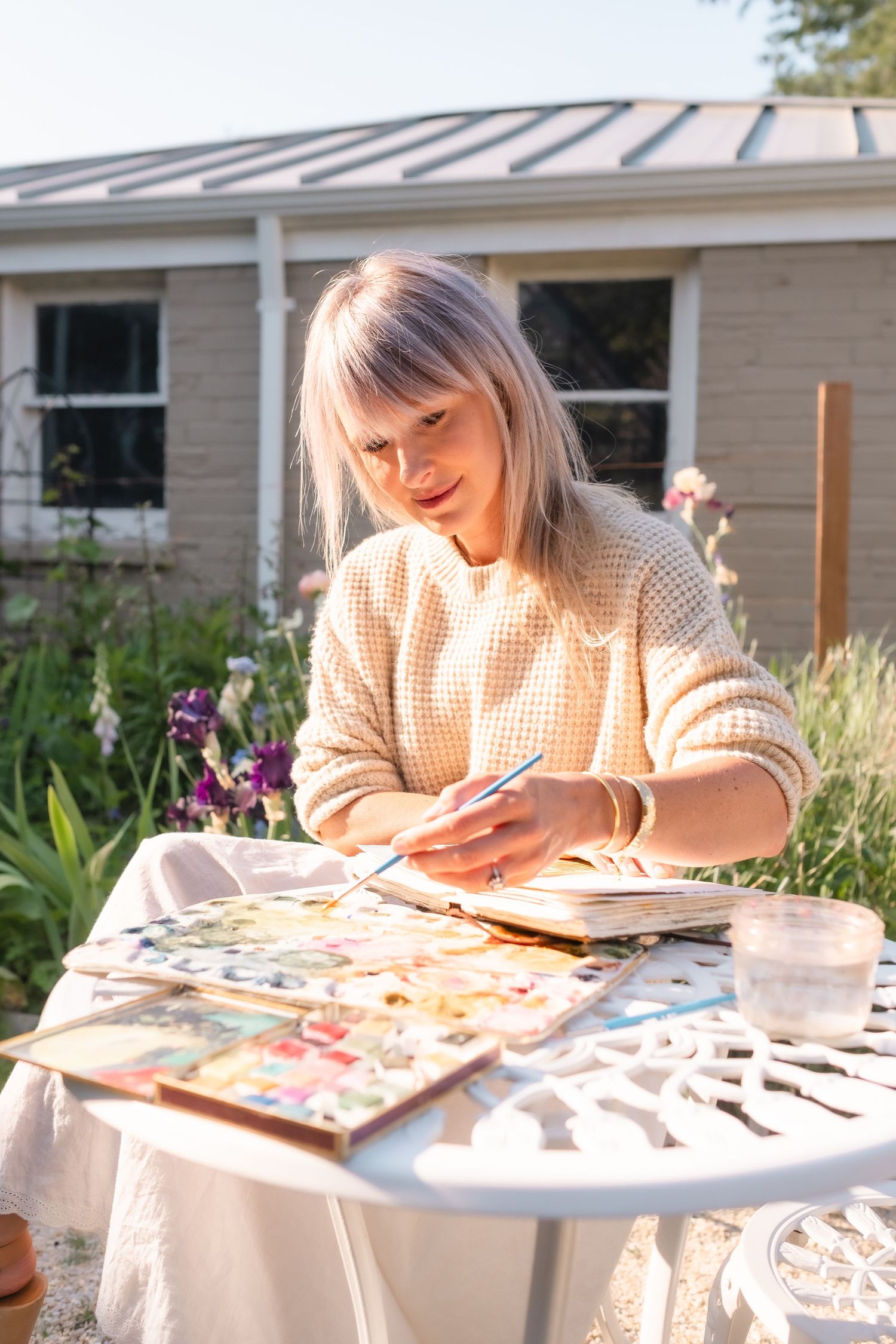 Woman painting watercolors outdoors at a white table in a sunlit garden near her home.
