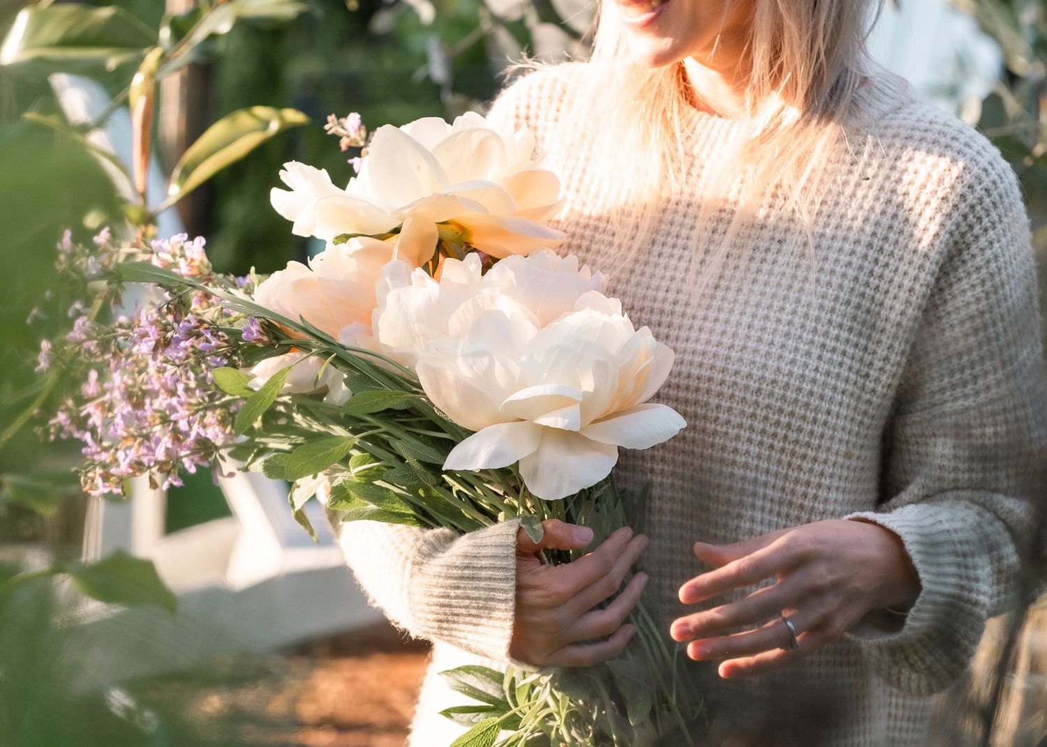 Woman in beige knit sweater holding white peony bouquet in garden sunlight.