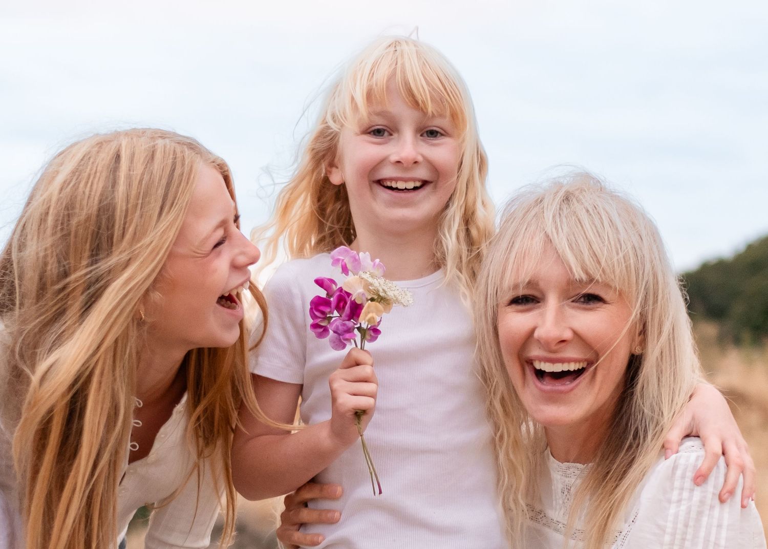 Three generations share joyful laughter together outdoors while holding pink flowers on a sunny day.