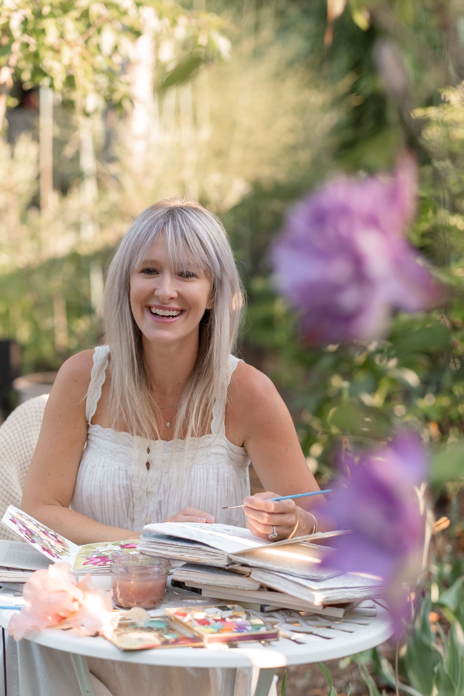 Smiling blonde woman sitting at outdoor table with books and art supplies surrounded by purple flowers.