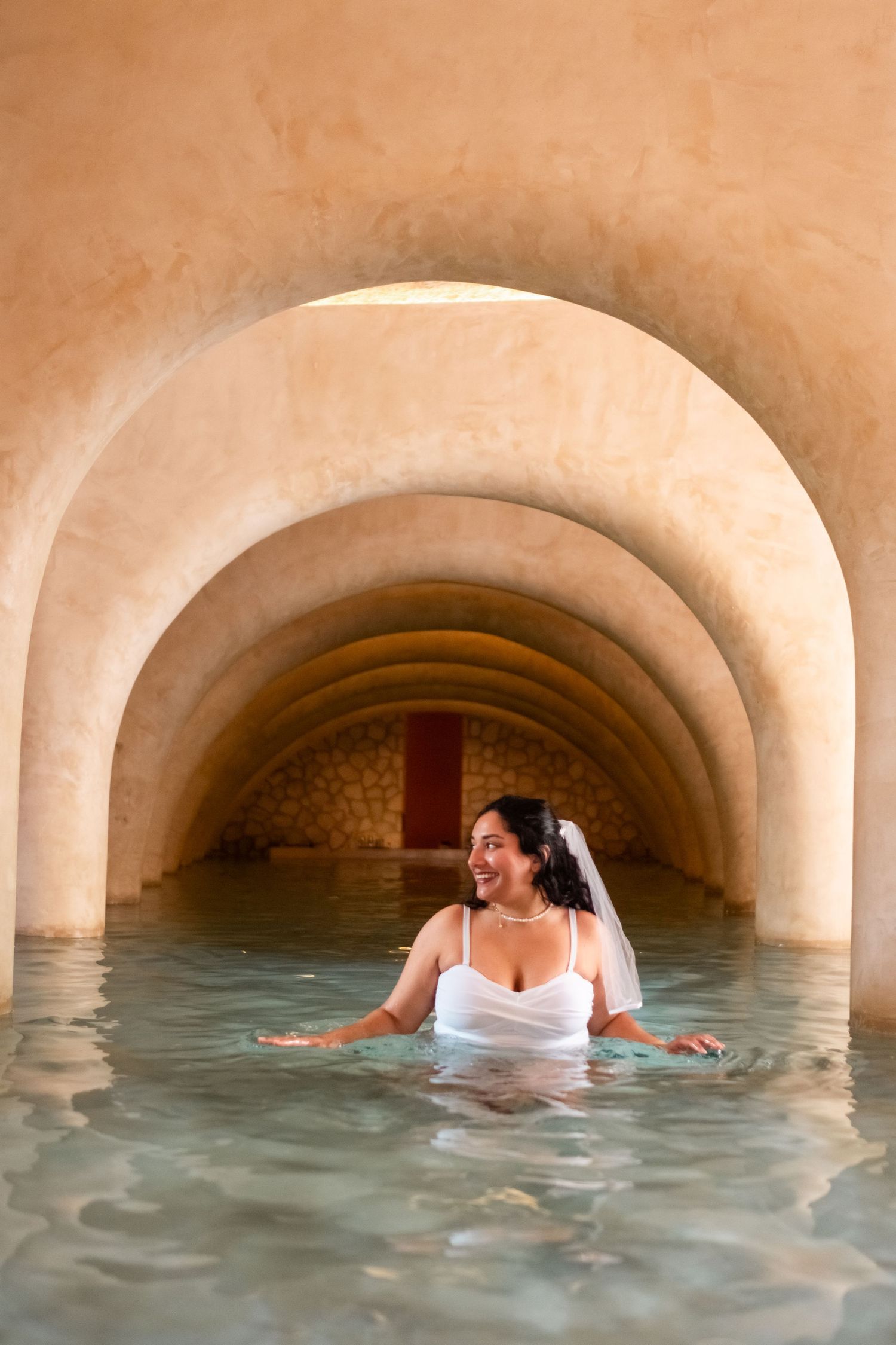 Smiling bride in white wedding dress and veil wading through water under arched stone tunnel ceiling.