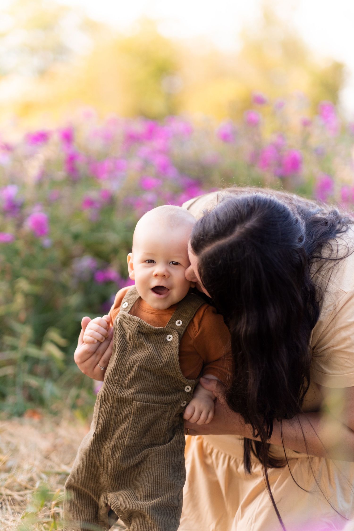 A baby wearing brown overalls stands in a garden with pink flowers blooming in the background during golden hour.