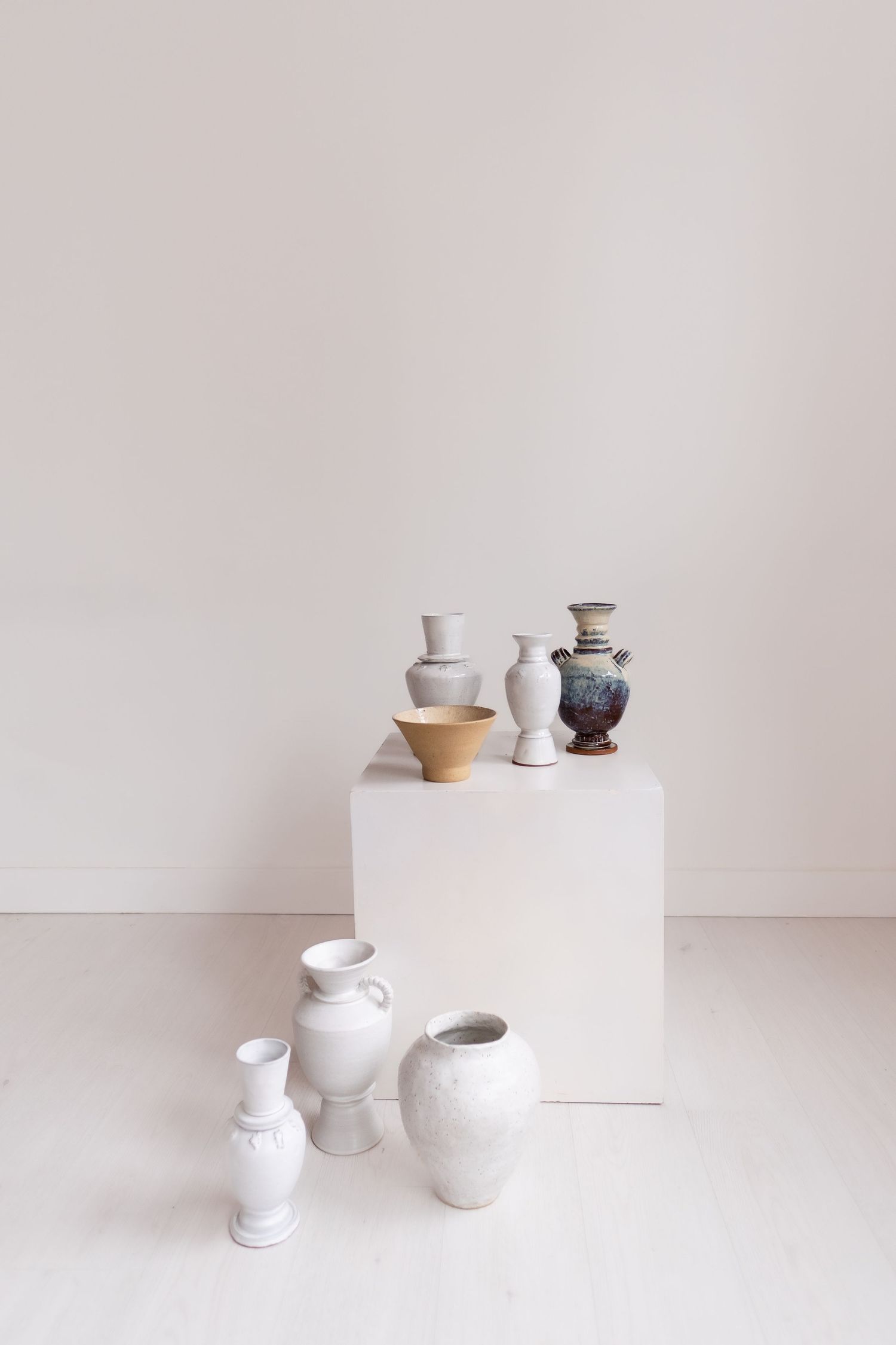 Minimalist still life composition with ceramic vases and pottery arranged on white pedestal against neutral background.