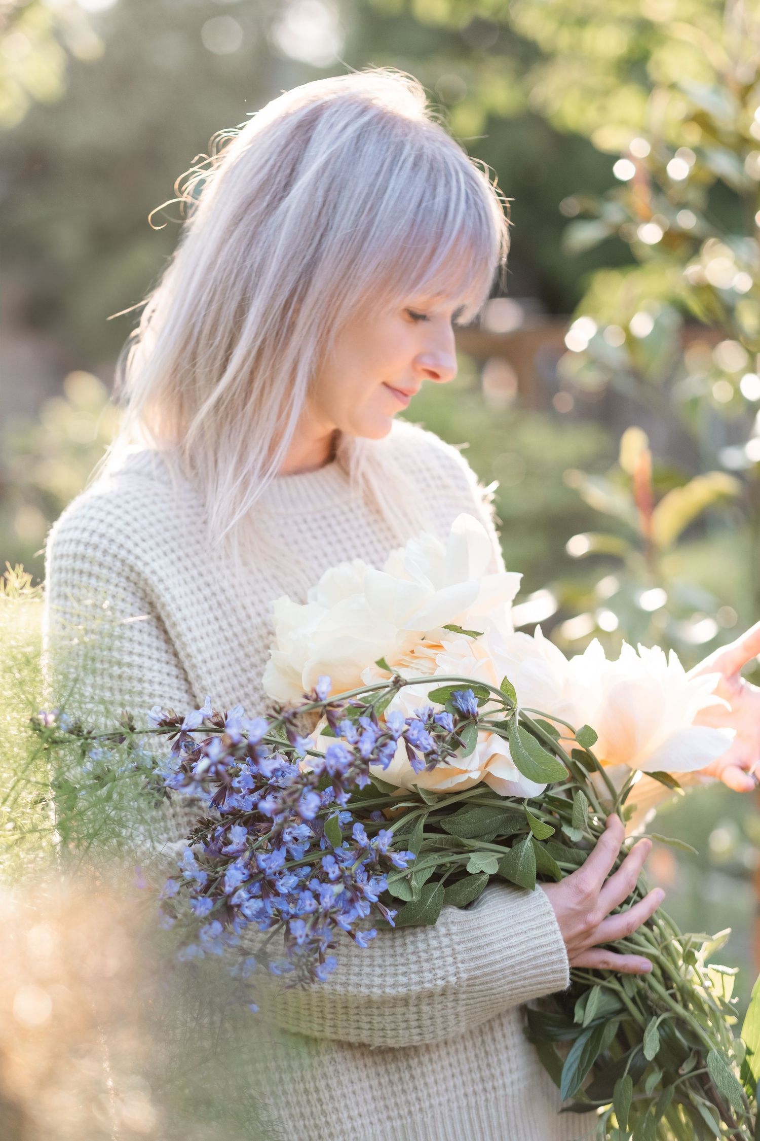 Blonde woman in beige waffle knit sweater holding a bouquet of white and blue flowers in sunlit garden.