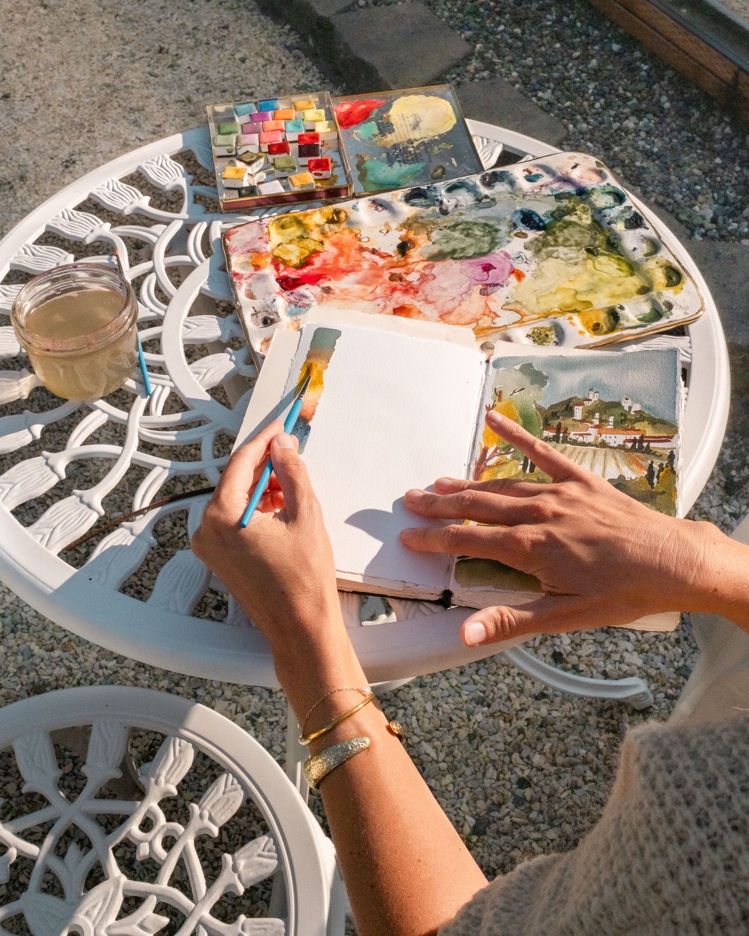 Hands painting watercolors in a sketchbook on a white ornate garden table with paint palettes.