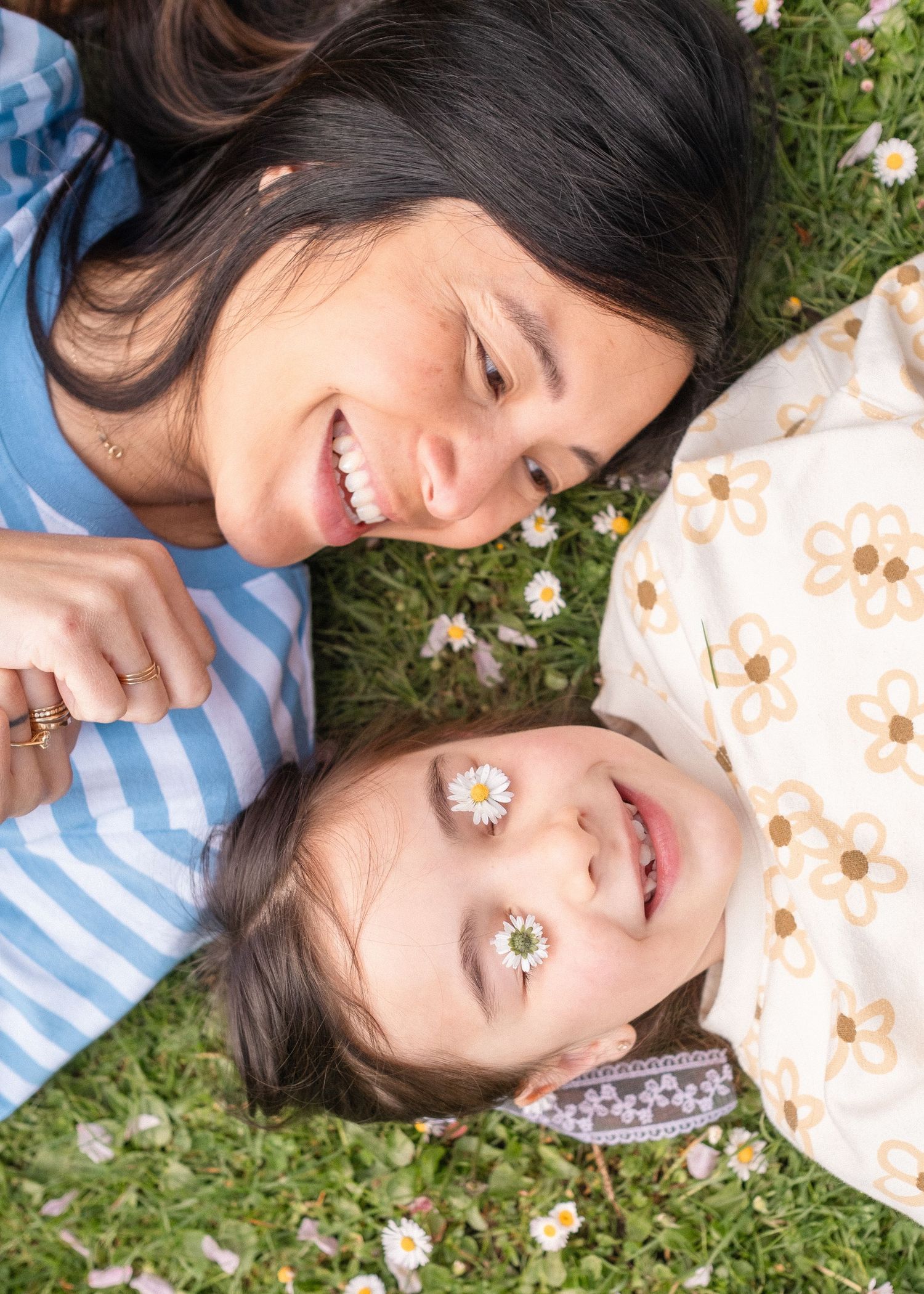 Two people lying on grass with daisies, sharing joyful laughter in a sunny garden setting.
