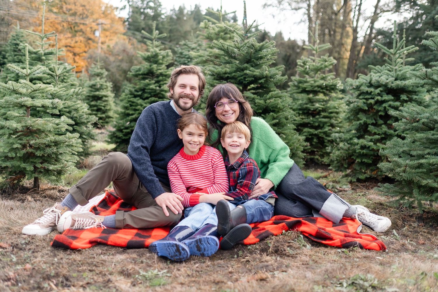 Family of four smiling together on a red plaid blanket at a Christmas tree farm surrounded by evergreen trees.