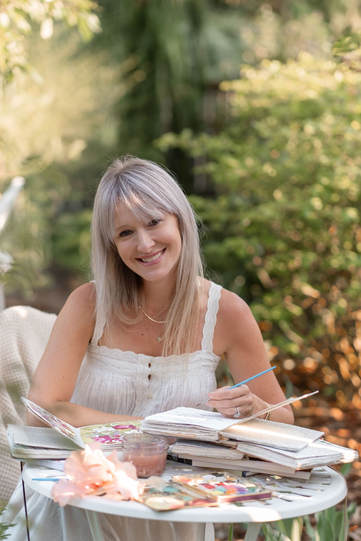 Smiling blonde woman painting in a sketchbook at an outdoor table surrounded by art supplies in a garden setting.