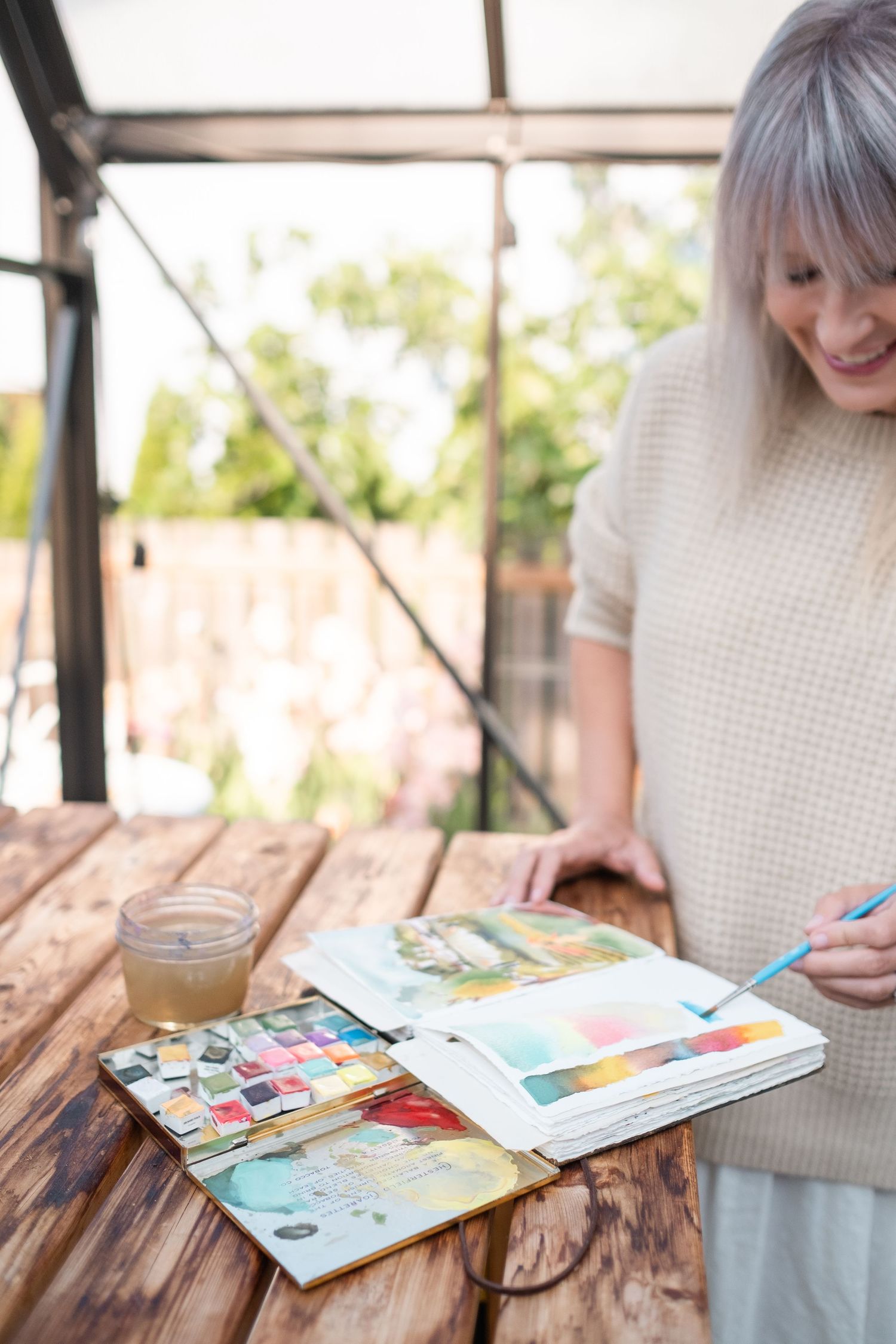 Woman painting in watercolor sketchbook at rustic wooden table inside a bright glass greenhouse with candle nearby.