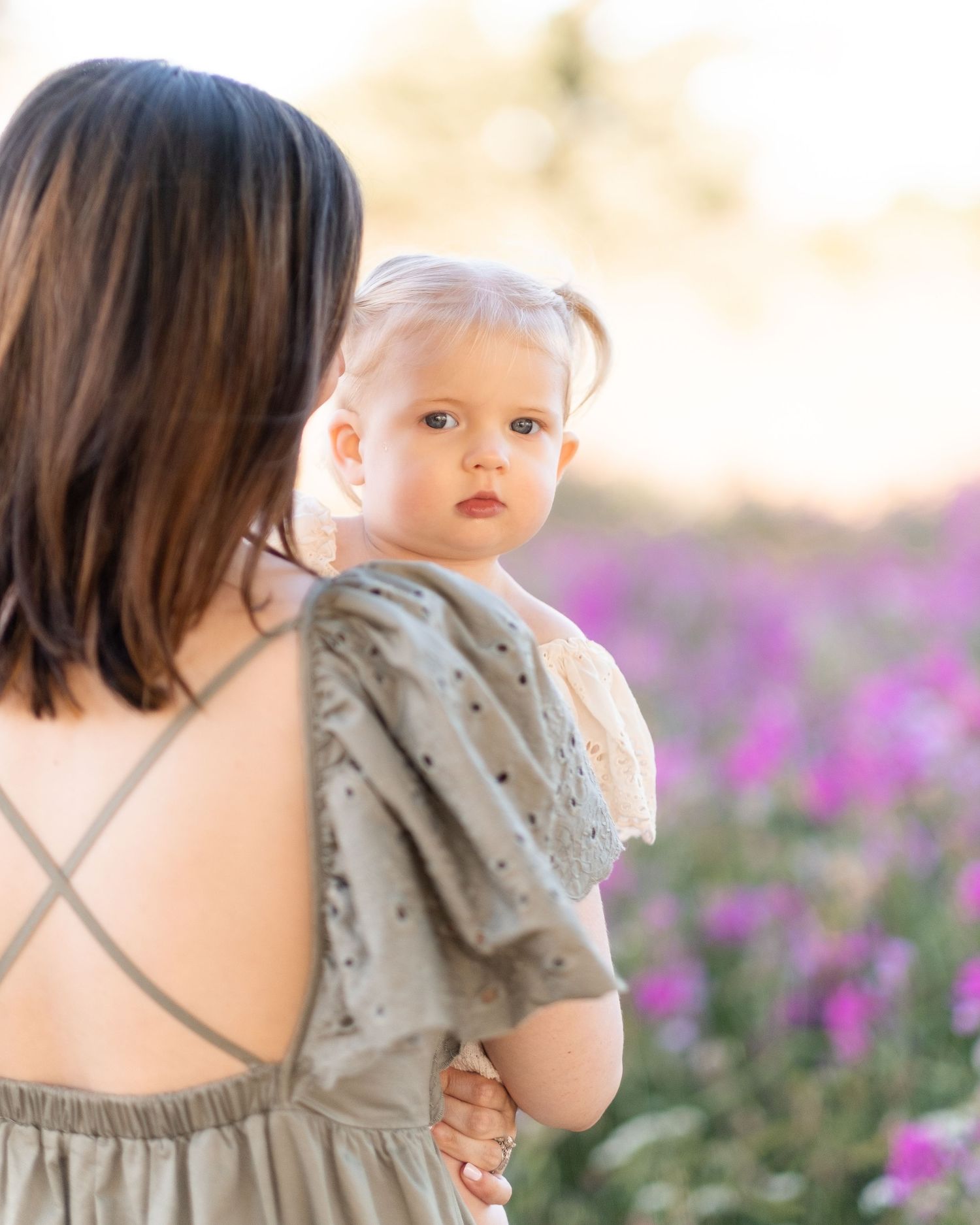 A baby in a light gray dress sits in a field of purple wildflowers during sunset.